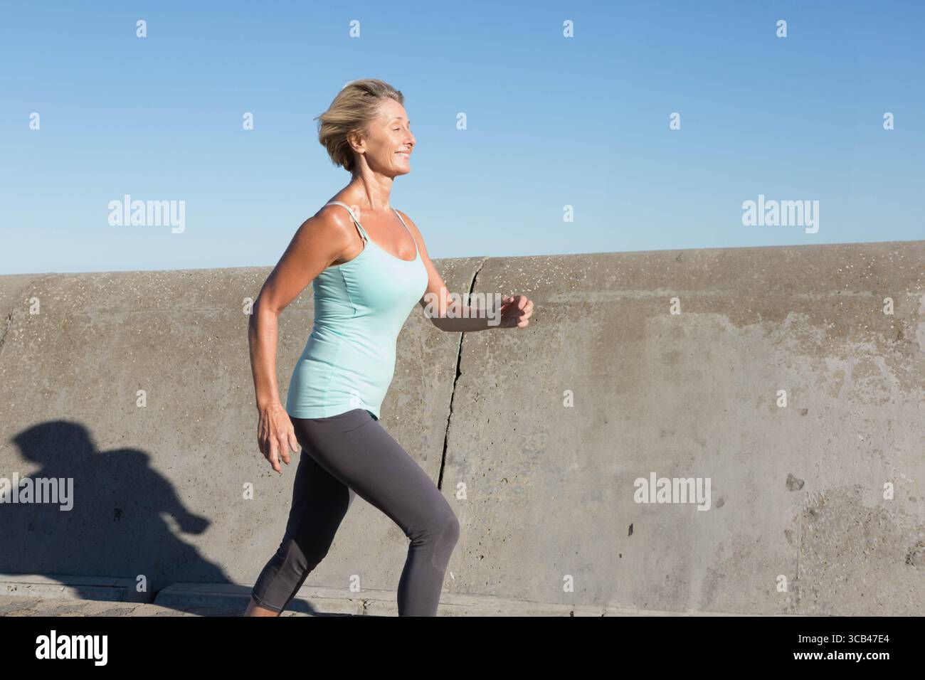 Seniorin joggt auf dem Gehweg an der Betonmauer in Sportbekleidung mit Sportschuhen, Kopierraum Stockfoto