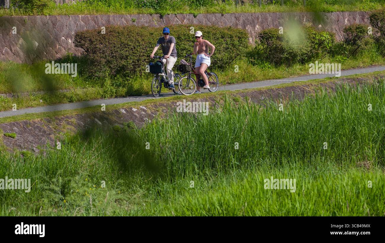 Zwei Personen radeln auf einem malerischen Naturpfad, umgeben von üppigem Grün unter der warmen Sonne, Kyoto, Japan Stockfoto