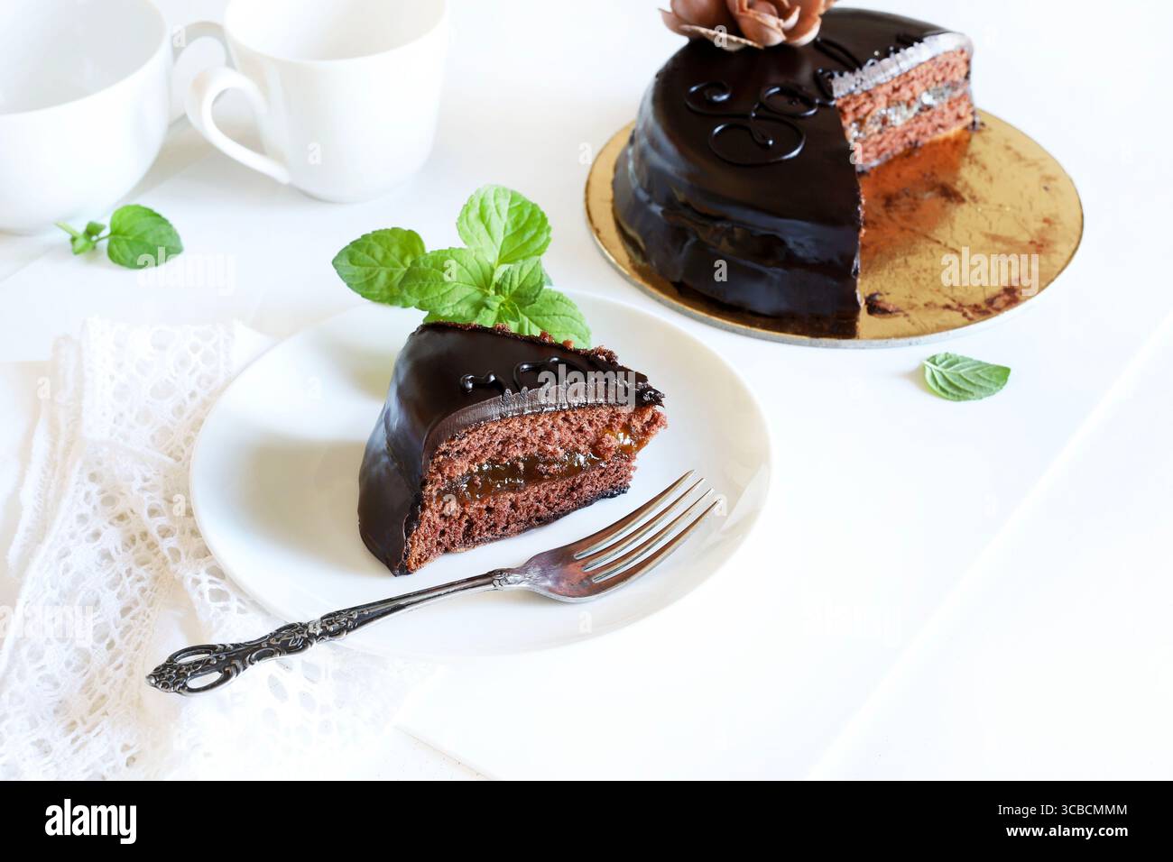 Sachertorte (Schokoladen-sacher-Kuchen) - Scheibe auf einem weißen Teller, frische Minzdekoration, ganzer Kuchen im Hintergrund. Traditionelles österreichisches Dessert. Stockfoto