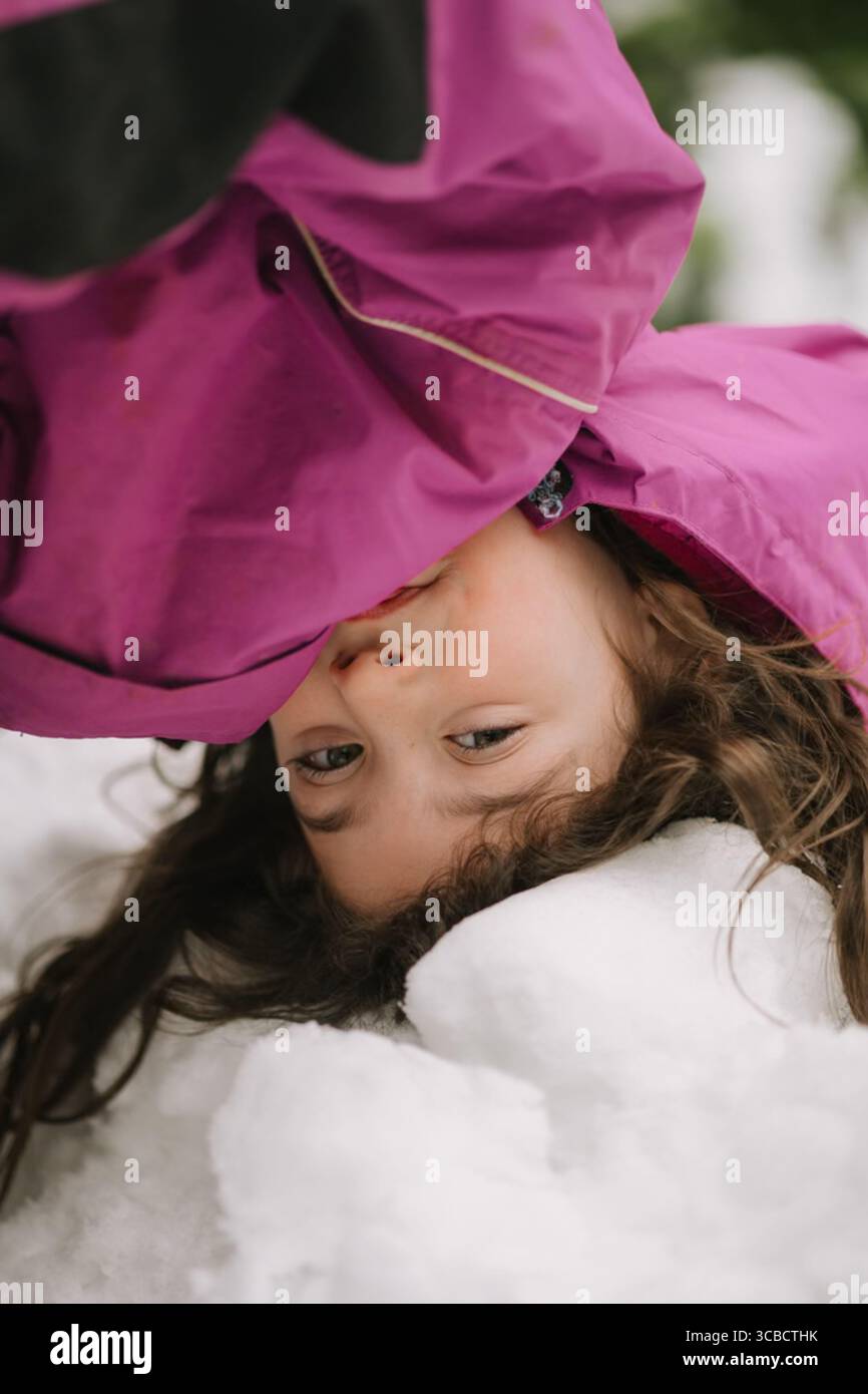 Kind in rosafarbener Jacke, das verspielt kopfüber im Schnee liegt Stockfoto
