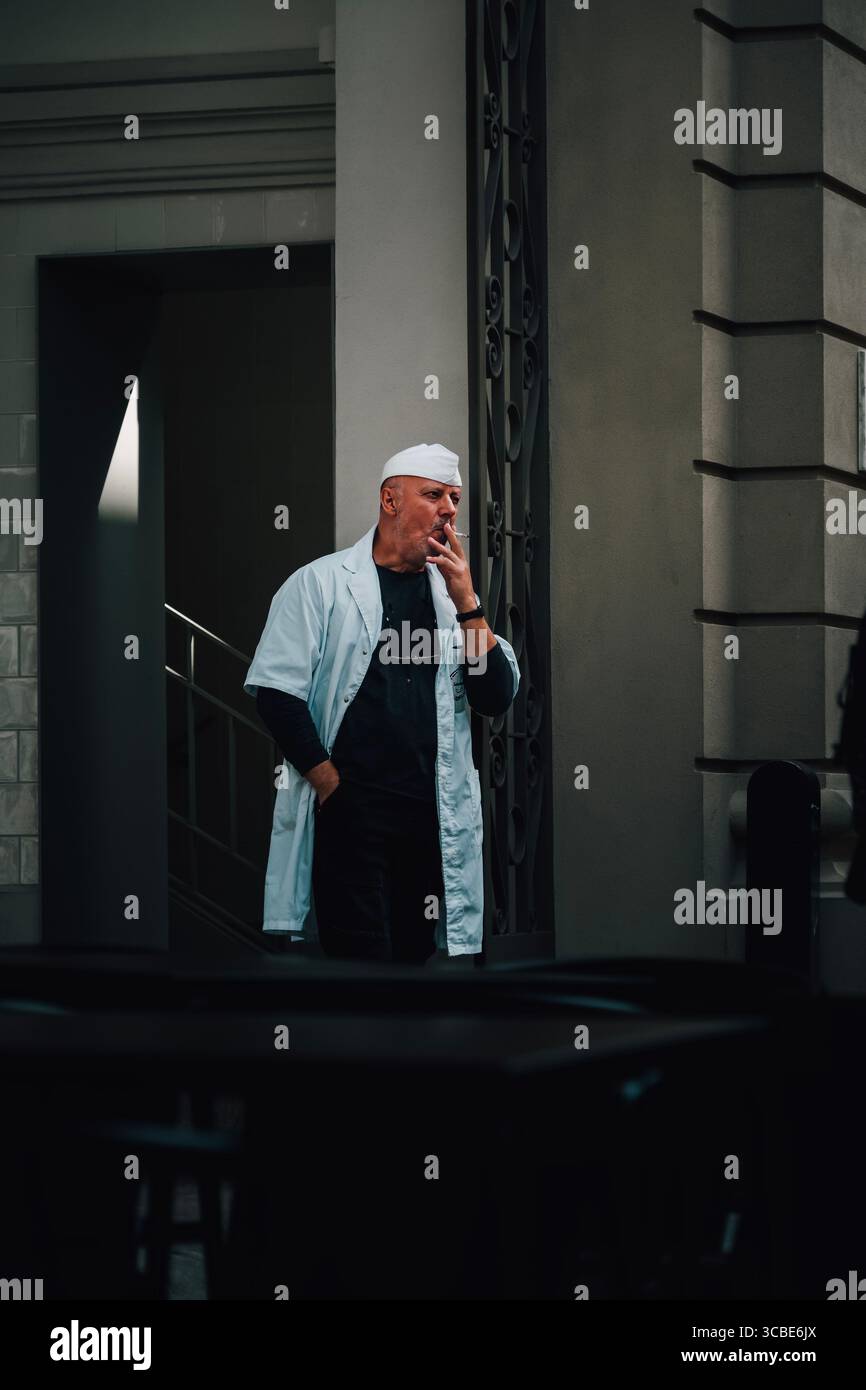 Mann in White Chef's Coat macht eine Rauchpause in einer urbanen Gasse von Porto. Portugiesisches Stadtleben, ungezwungener Moment, wenig Licht, europäisches Straßenbild Stockfoto