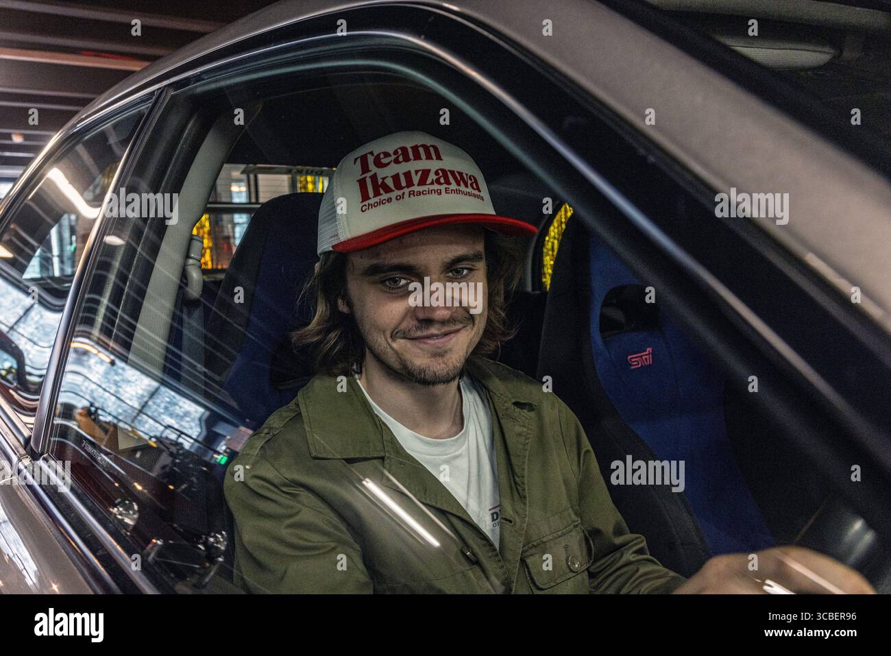 Junger Mann mit einer Team Ikuzawa Rennmütze hinter dem Lenkrad eines Autos, der die Motorsporttradition des legendären japanischen Rennteams demonstriert. Stockfoto