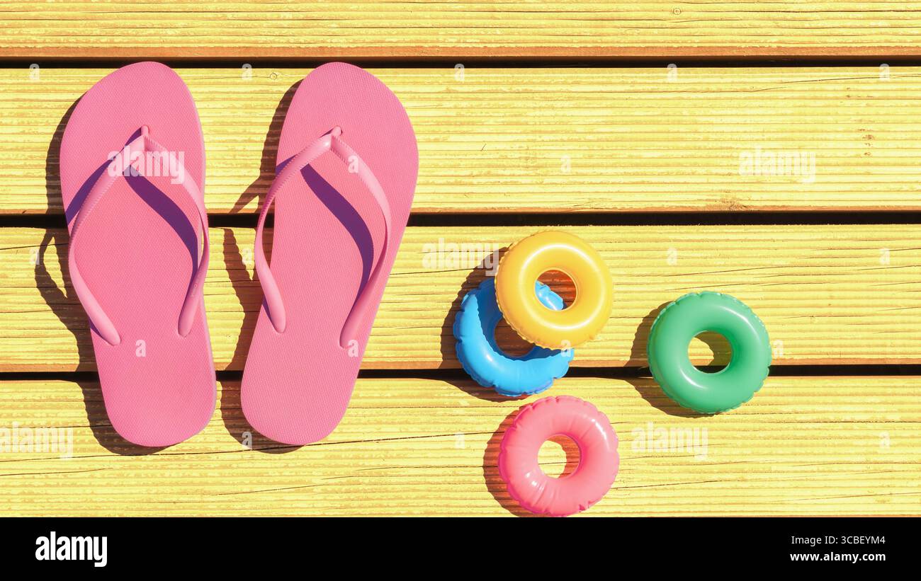 Leuchtend rosa Flipflops ruhen auf gelben Holzterrassen neben farbenfrohen Schwimmerspielzeugen. Die lebhafte Szene fängt einen sonnigen Tag ein, perfekt zum Entspannen Stockfoto