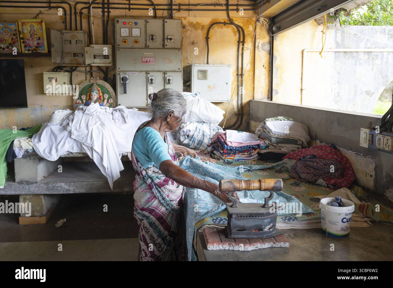 Ältere Frau in traditionellen Kleiderbügeln mit einem antiken Holzkohleeisen, Dhobi Ghat, Freiluftwäscherei in Kochi, Indien, 20.03.2025 Stockfoto
