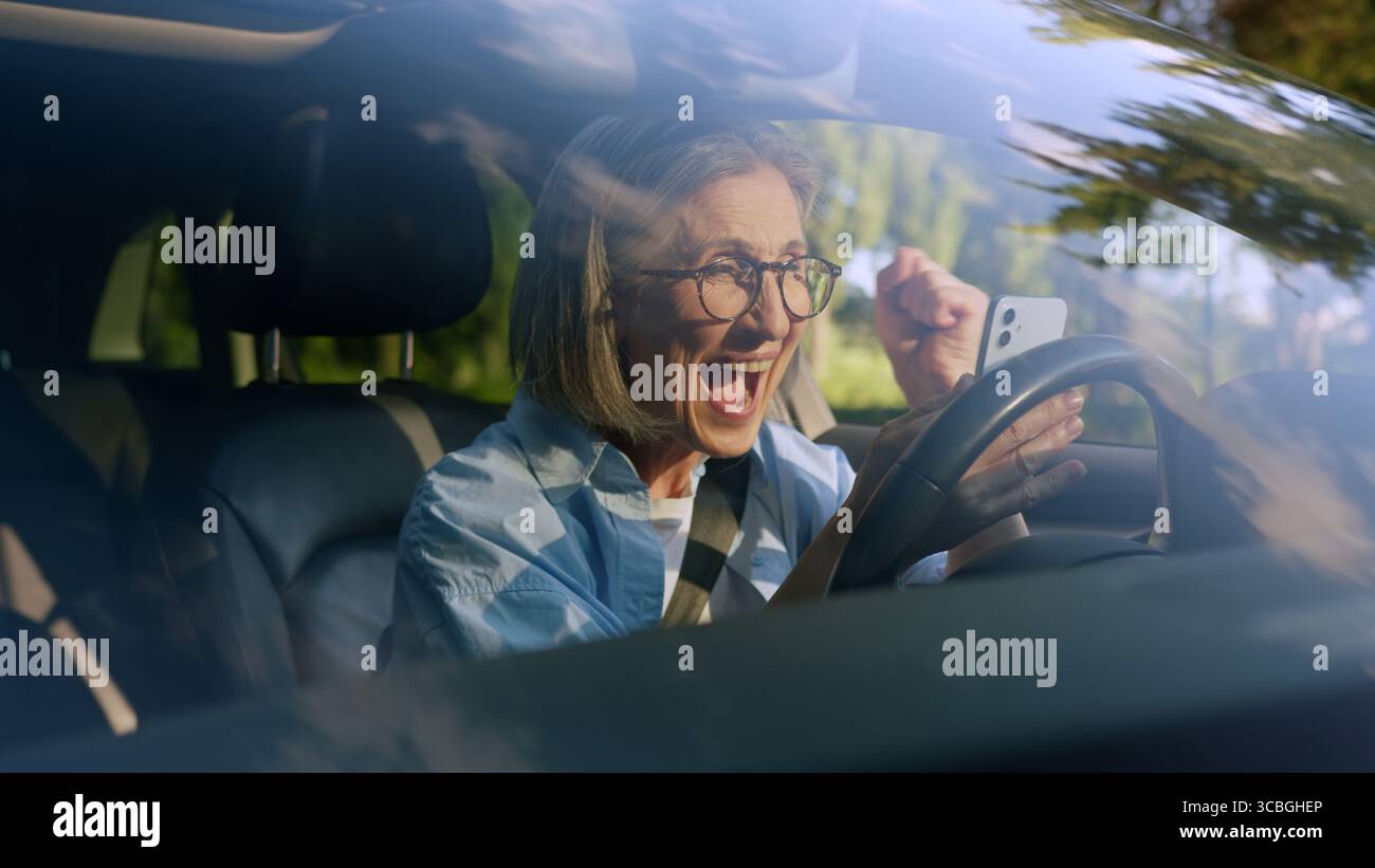 Aufgeregt schockiert überrascht kaukasische ältere ältere Frau weibliche ältere Fahrer Auto Besitzer im Auto mit Handy-App gewinnen lesen großartig Stockfoto