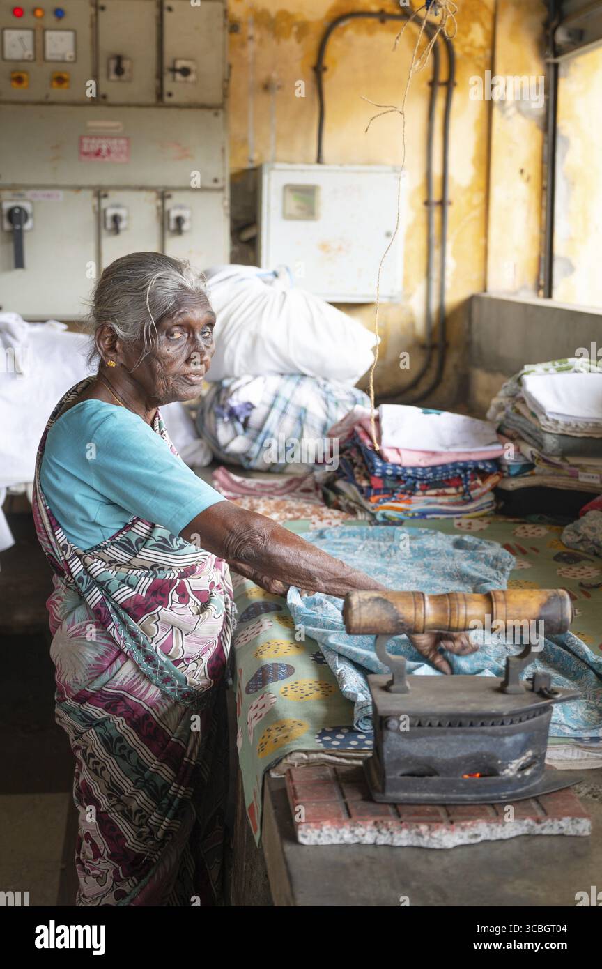 Ältere Frau in traditionellen Kleiderbügeln mit einem antiken Holzkohleeisen, Dhobi Ghat, Freiluftwäscherei in Kochi, Indien, 20.03.2025 Stockfoto