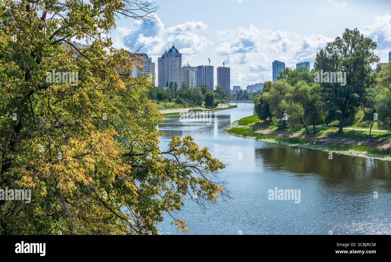 Üppige Bäume säumen den Fluss Dnipro, während er sich durch Rusaniwka in Kiew, Ukraine, schlängelt, mit modernen Wohngebäuden im Hintergrund darunter Stockfoto