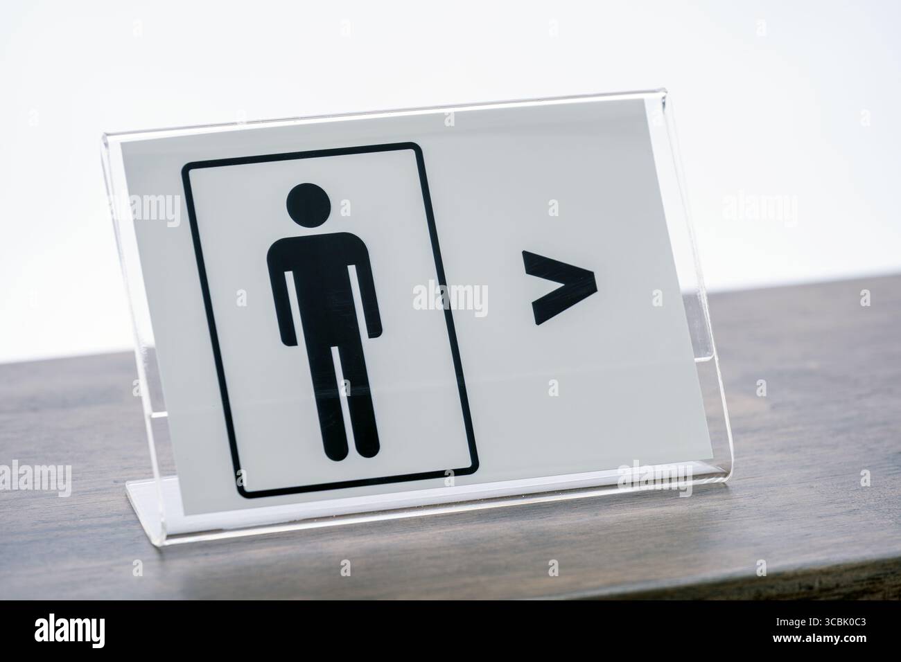 Ein klares Acrylschild mit männlichem WC-Symbol und Richtungspfeil befindet sich auf einer Holztheke und zeigt den Standort der männlichen Toilette an Stockfoto