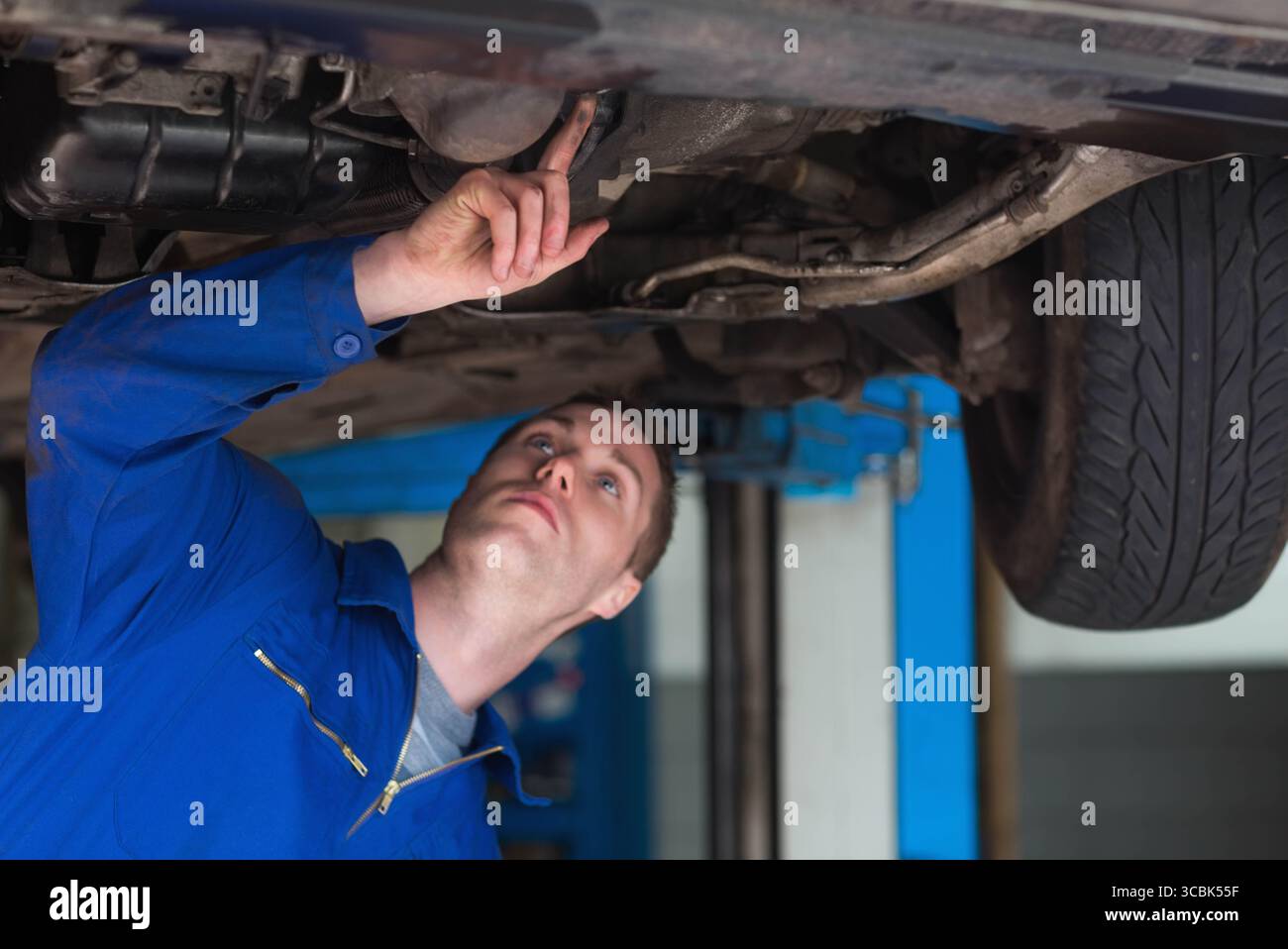 Männlicher Kfz-Techniker, der auf den Auspuff zeigt, untersuchte das Laufwerk auf dem Aufzug in der Werkstatt, Kopierraum. Mechaniker, Wartung, Inspektion, Werkstatt, Prec Stockfoto