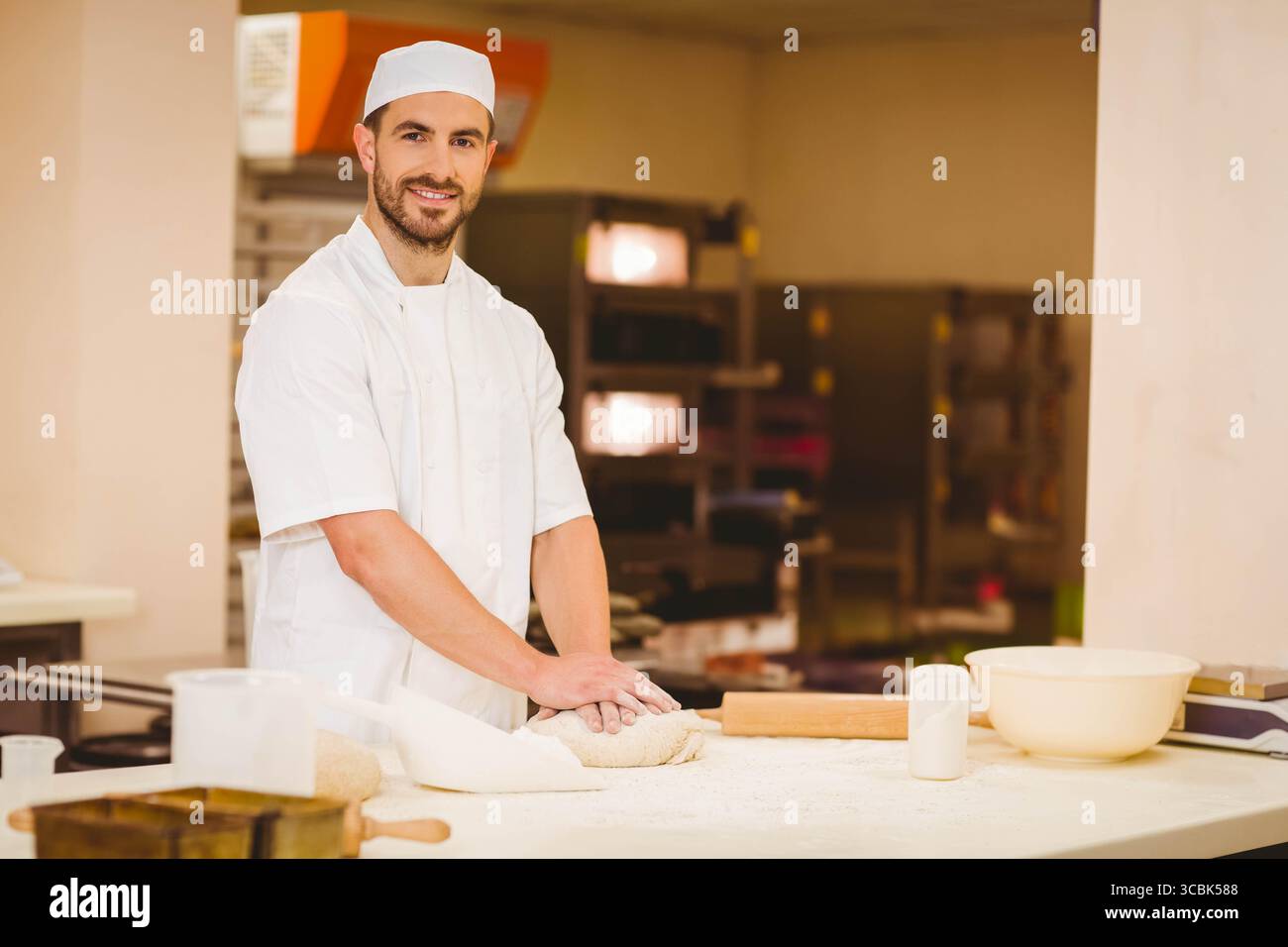Männlicher Bäcker mit weißem, gleichmäßigem Knetteig auf bemehlter Arbeitsplatte in der Bäckerei mit Nudelnadel. Gebäck, Küche, Kunsthandwerk, Edelstahl, Kunsthandwerk Stockfoto