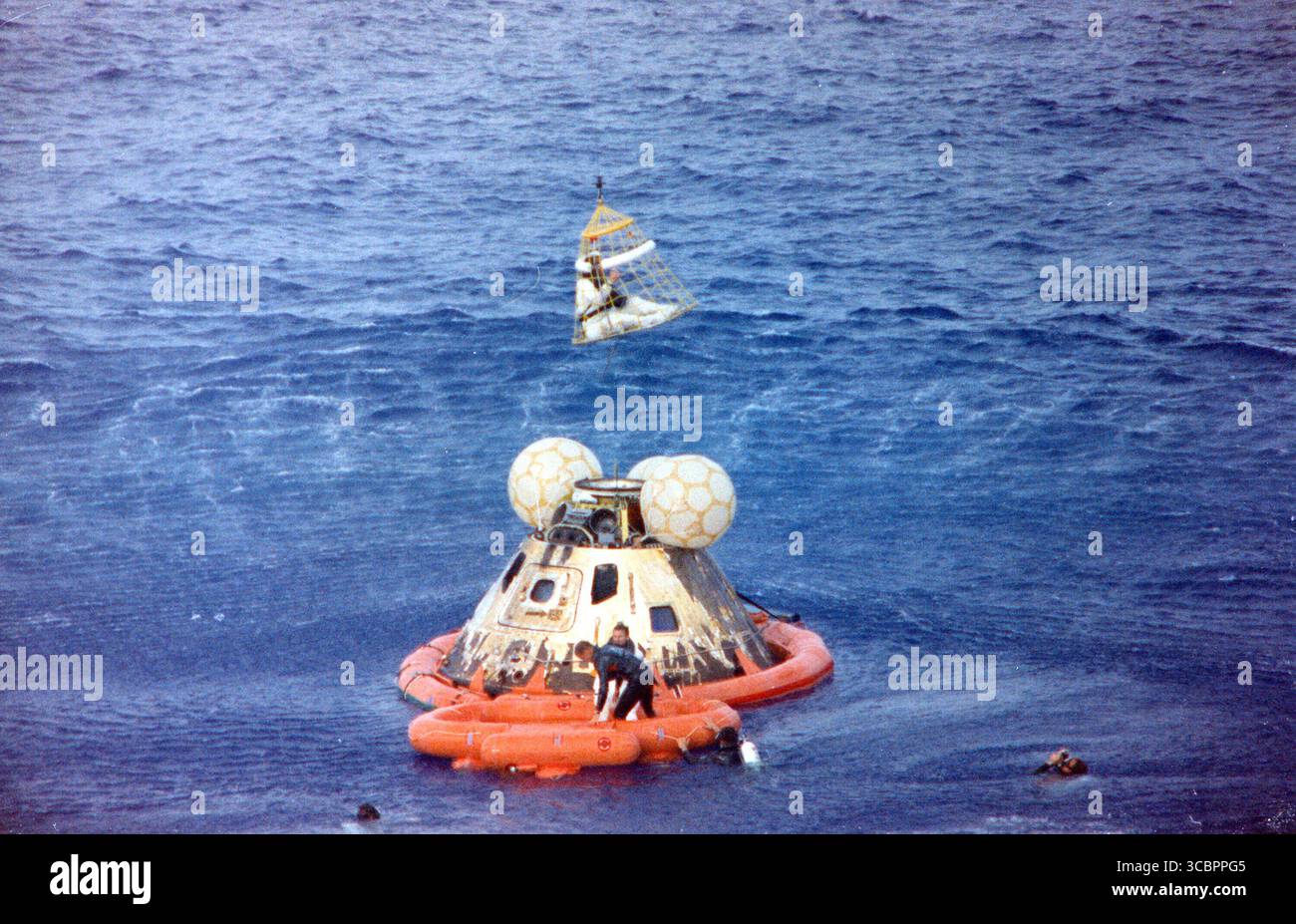 Astronaut John L. Swigert Jr., Pilot des Kommandomoduls, wird in einem Billy Pugh Hubschrauberrettungsnetz an Bord eines Hubschraubers gehoben, während Astronaut James A. Lovell Jr., Commander, auf ihn wartet. Astronaut Fred W. Haise Jr., Pilot des Mondmoduls, ist bereits an Bord des Hubschraubers. Im Rettungsfloß mit Lovell und im Wasser befinden sich mehrere Unterwasser-Abbruchteams der U.S. Navy, die bei den Bergungseinsätzen mitgeholfen haben. Die Besatzung wurde auf die US-amerikanische Insel Iwo Jima gebracht, einige Minuten nachdem die Apollo 13 am 17. April 1970 um 12:01:44 Uhr CST niedergespritzt war Stockfoto