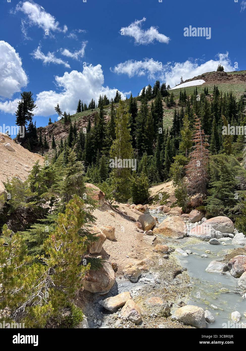 Geothermischer Fluss und felsiges Gelände in Bumpass Hell im Lassen Volcanic National Park, Kalifornien, mit bewaldeten Hängen und hellem Sommerhimmel. - Smartphone-aufgenommenes Stockfoto
