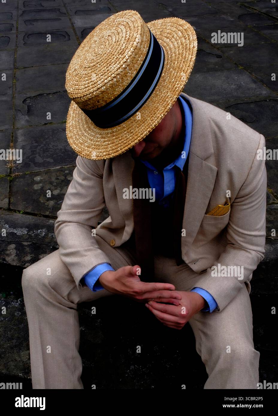 Ein Mann mit Strohhut, Anzug und Krawatte sitzt auf einer Steinoberfläche und blickt auf seine Hände. Stockfoto