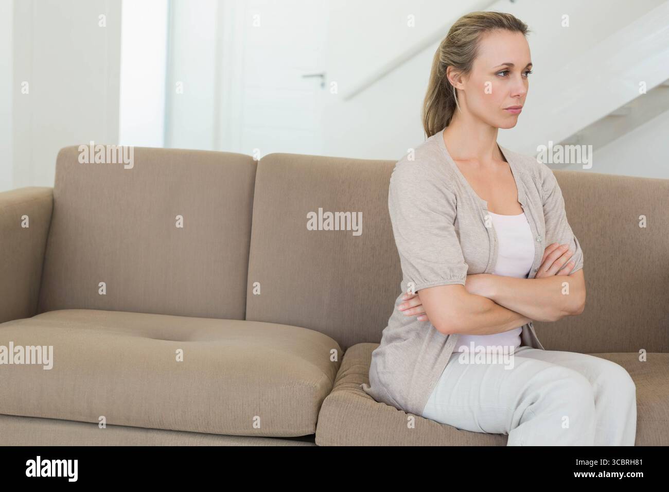 Frau mittleren Alters, die mit gekreuzten Armen auf beigem Sofa sitzt, im Wohnzimmer bei Treppe, Kopierraum Stockfoto