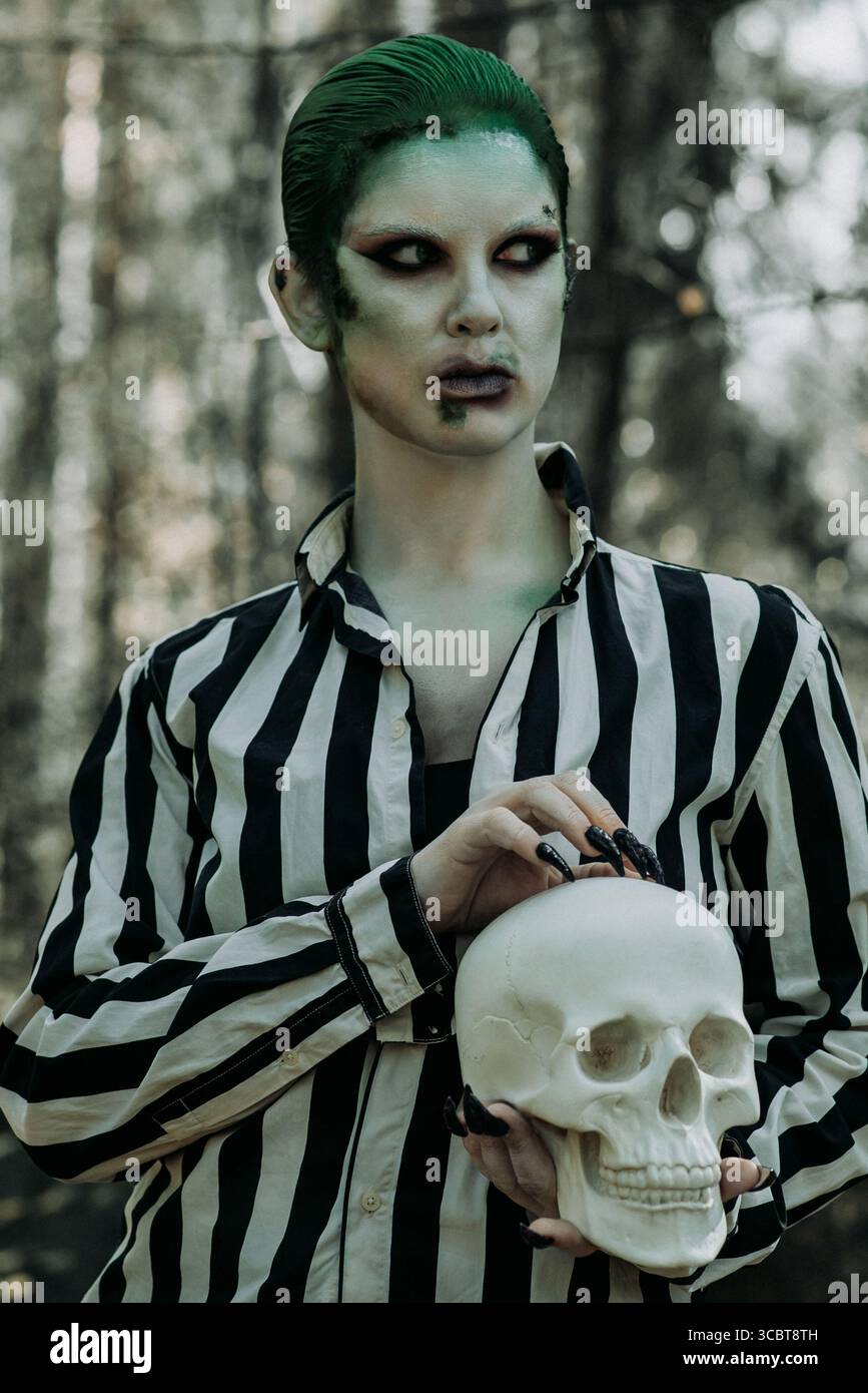 Fantasy-Charakter in grüner Gesichtsfarbe und gestreiftem Hemd mit menschlicher Schädelstütze, halloween inspiriertes Porträt in Waldlage Stockfoto