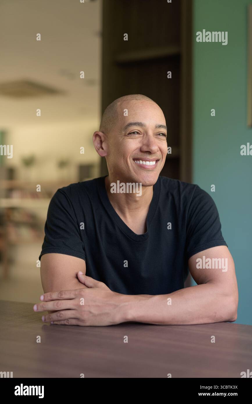 Glücklicher, kahlköpfiger hispanic-Mann in lässigem schwarzem T-Shirt, der in der Bibliothek sitzt und lächelt, während er aus dem Fenster blickt. Heller, moderner Innenraum mit Bücherregalen und Stockfoto