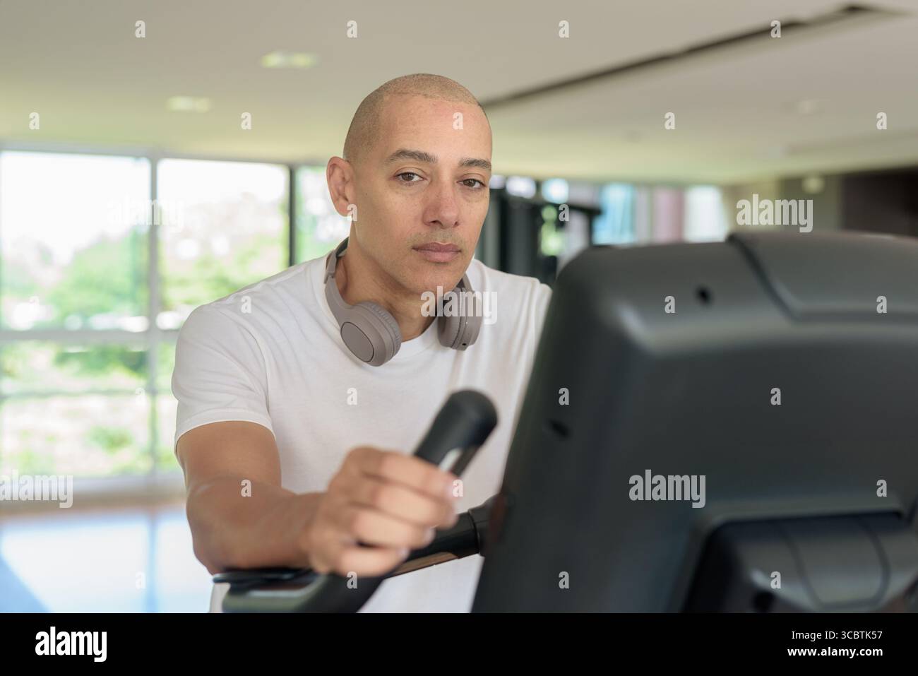 Glatze Hispanic Mann, der auf einem Heimtrainer in einem modernen Fitnessstudio trainiert. Fokussiertes Fitness- und Workout-Konzept, das Kraft, Herz-Kreislauf und Motive zeigt Stockfoto
