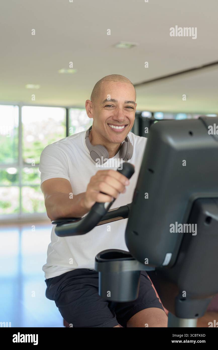 Glatze Hispanic Mann, der auf einem Heimtrainer in einem modernen Fitnessstudio trainiert. Fokussiertes Fitness- und Workout-Konzept, das Kraft, Herz-Kreislauf und Motive zeigt Stockfoto