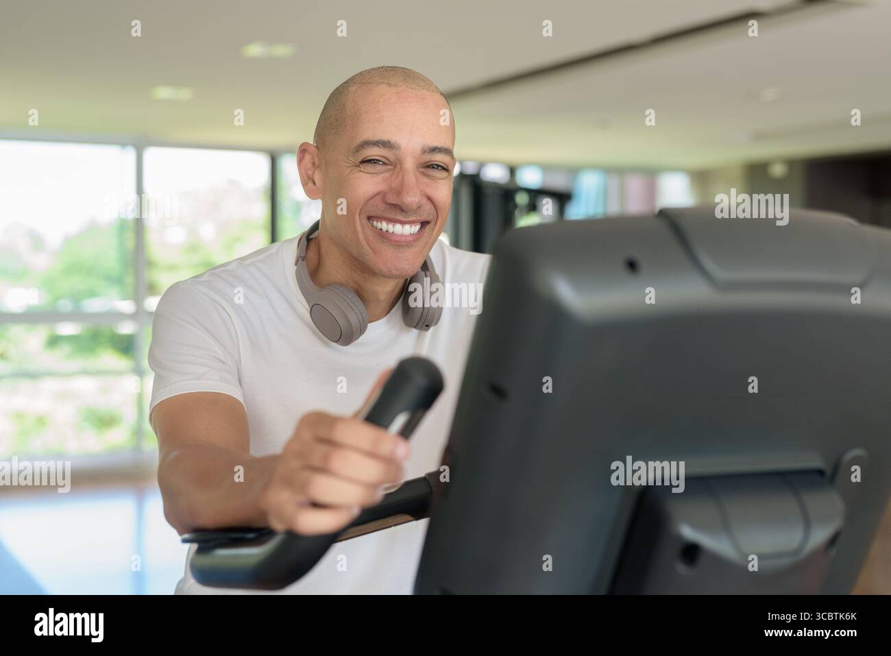 Glatze Hispanic Mann, der auf einem Heimtrainer in einem modernen Fitnessstudio trainiert. Fokussiertes Fitness- und Workout-Konzept, das Kraft, Herz-Kreislauf und Motive zeigt Stockfoto
