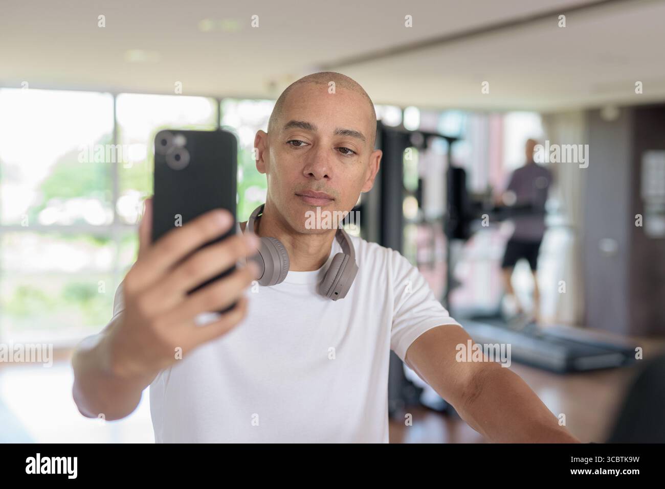 Kahler hispanischer Mann, der auf einem Heimtrainer in einem modernen Fitnessstudio trainiert, während er telefoniert. Fokussiertes Fitness- und Workout-Konzept, das Stärke zeigt, Stockfoto