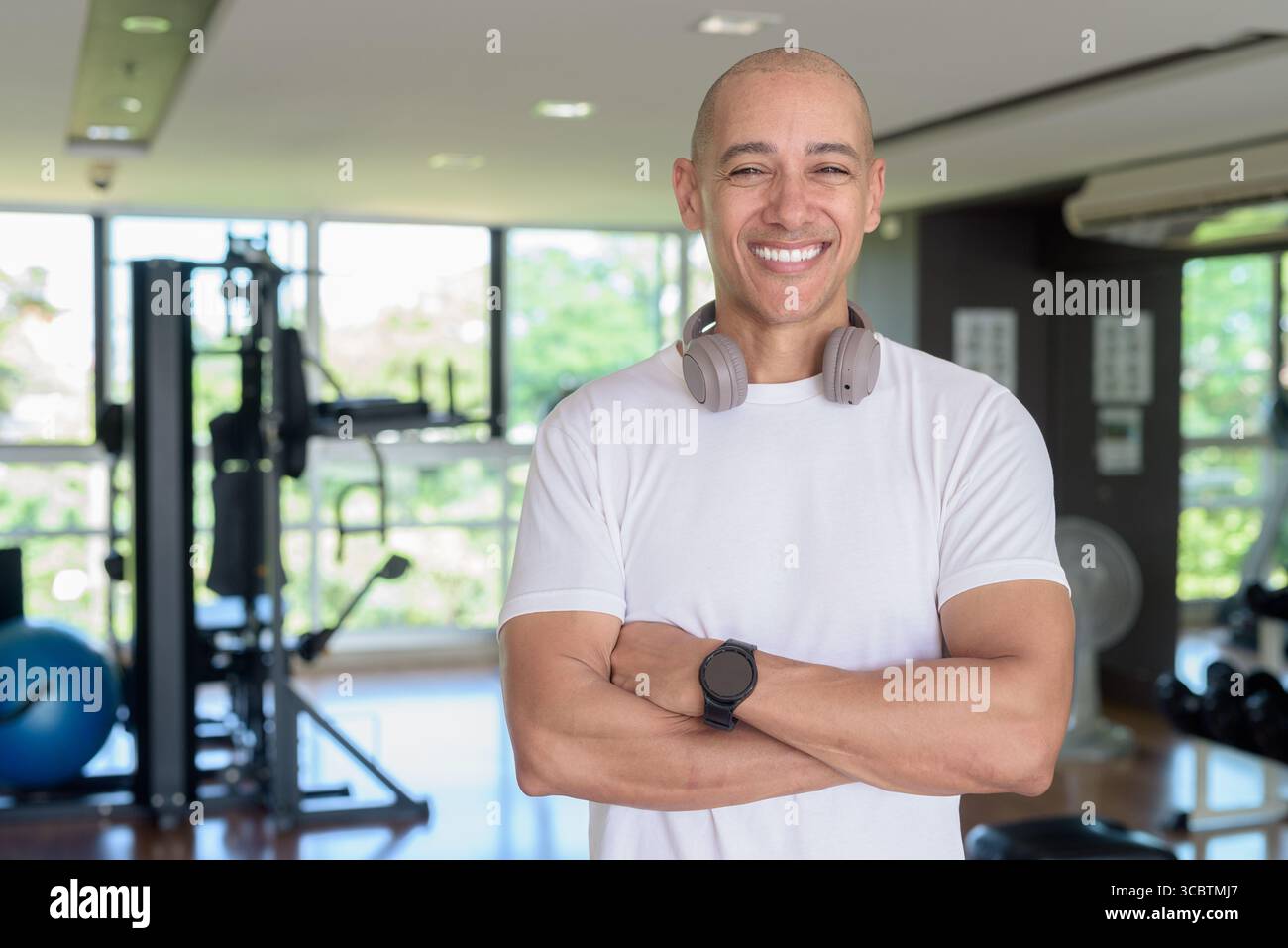 Glatze Latino Mann, der in einem modernen Fitnessstudio mit selbstbewusstem Ausdruck steht. Das Porträtieren von Wellness, Trainingsdisziplin und einem starken gesunden Lebensstil in indo Stockfoto
