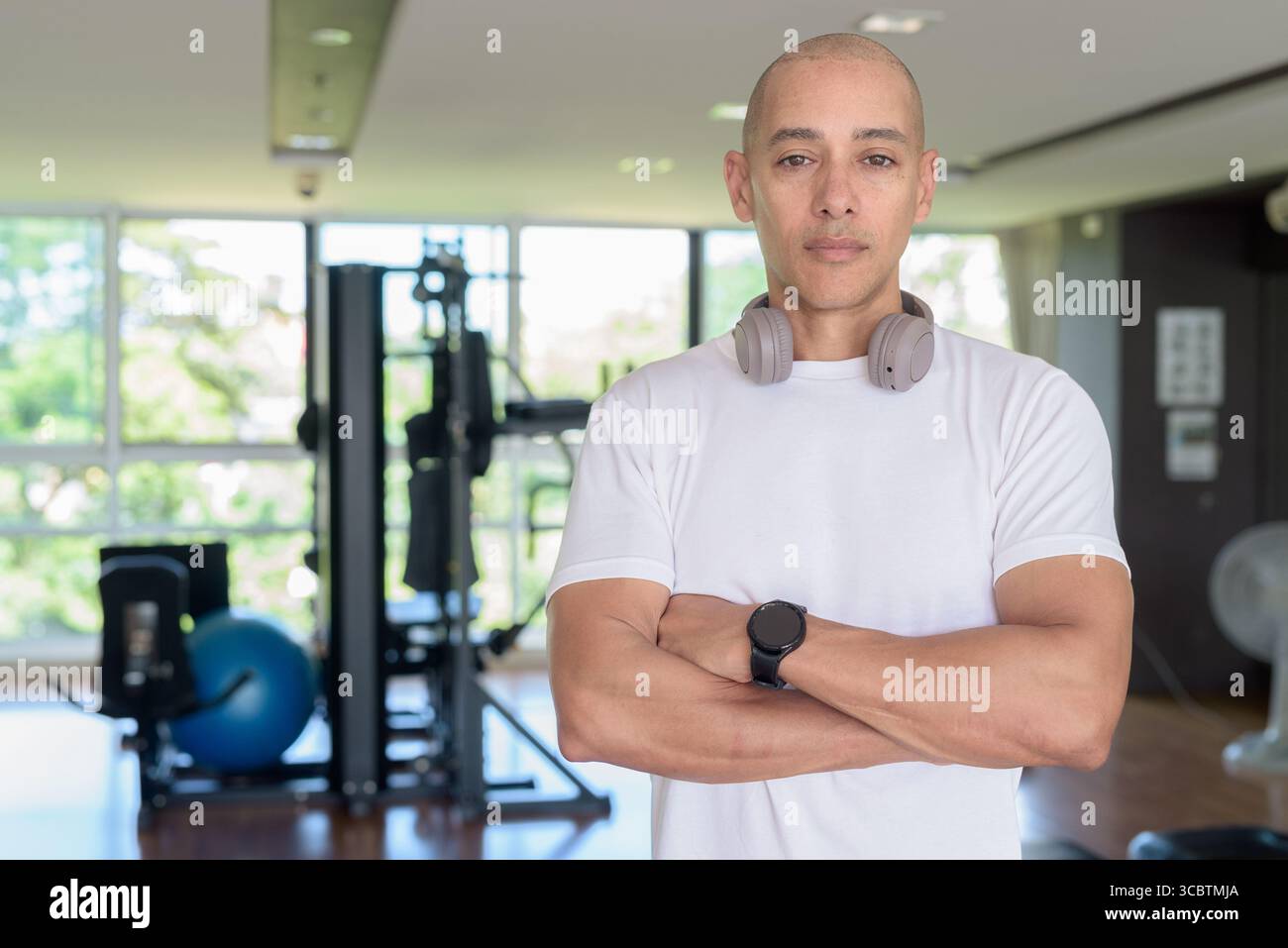 Glatze Latino Mann, der in einem modernen Fitnessstudio mit selbstbewusstem Ausdruck steht. Das Porträtieren von Wellness, Trainingsdisziplin und einem starken gesunden Lebensstil in indo Stockfoto