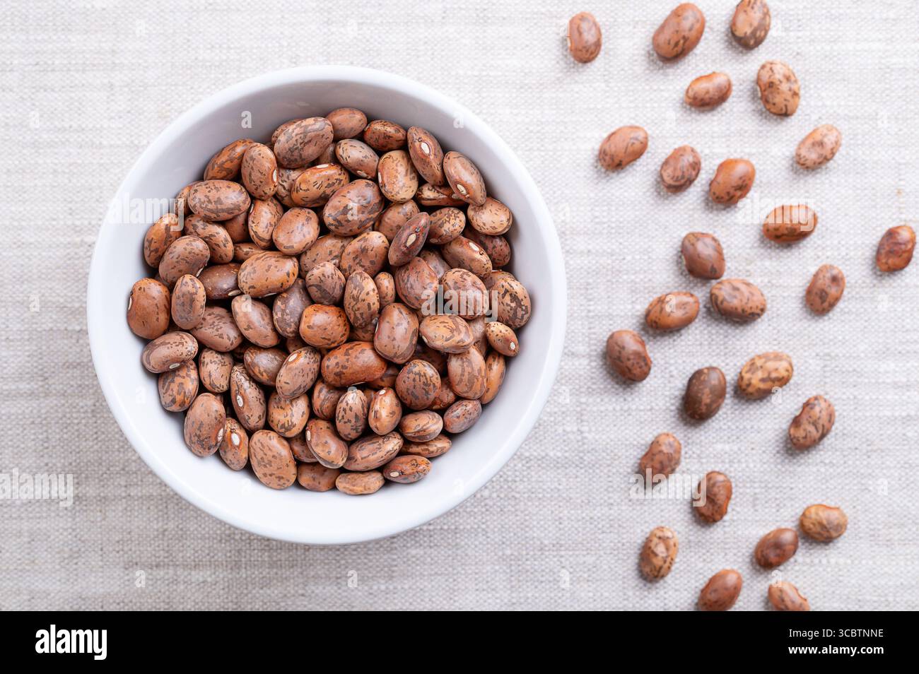 Pinto-Bohnen, in einer weißen Schüssel, auf Leinenstoff. Getrocknete Sorte gewöhnlicher Bohnen, Phaseolus vulgaris. Bohnenart, die braun mit rötlich-braunen Flecken ist. Stockfoto