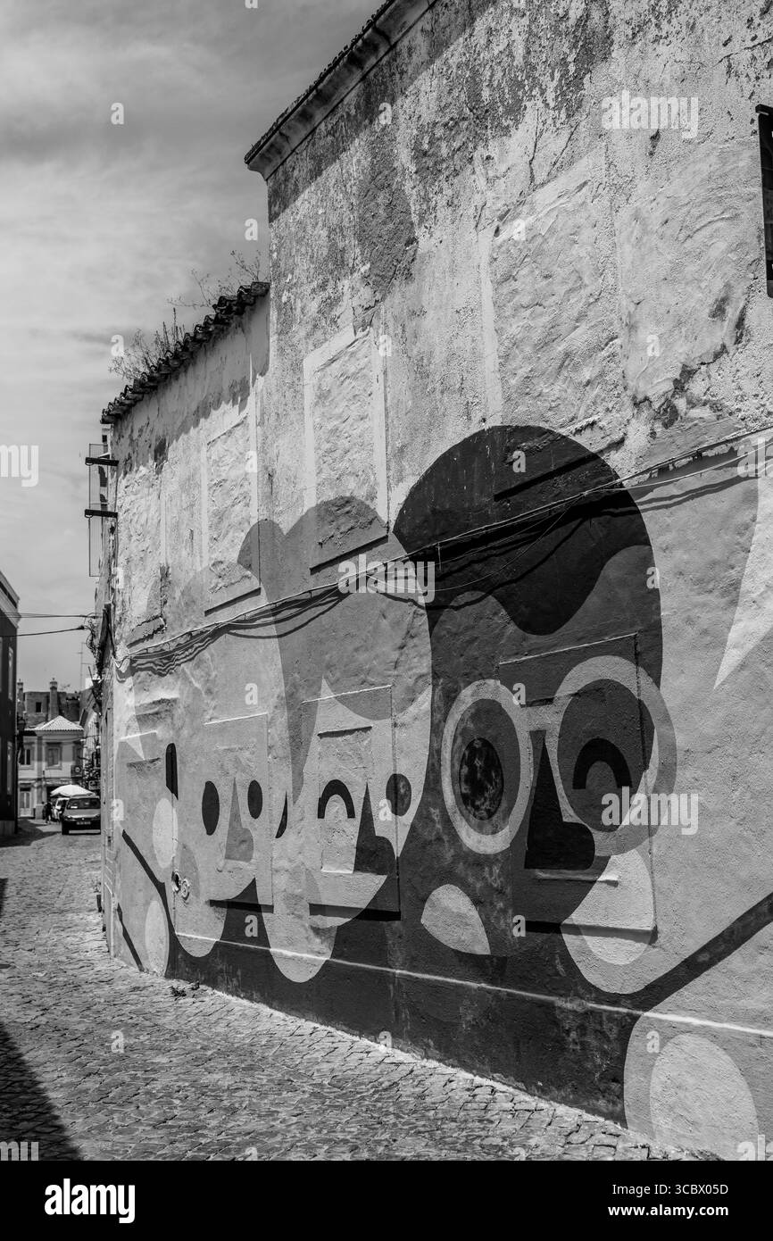 Algarve, Portugal – 2025.06.28 Ein lebendiges Wandbild mit Gesichtern im Cartoon-Stil, die auf einer verwitterten Wand in einer ruhigen Kopfsteinpflasterstraße gemalt sind Stockfoto