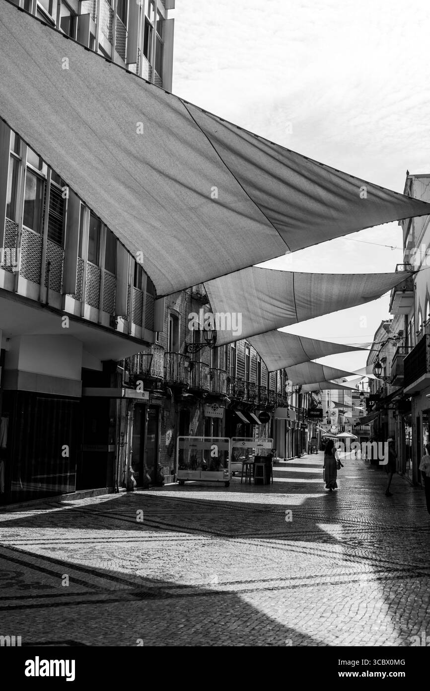 Algarve, Portugal – 2025.06.28 Eine bezaubernde, sonnendurchflutete Straße mit traditionellem Mosaikpflaster, Sonnenschirmen aus Stoff und Vintage-Balkonen in einem ruhigen Einkaufsviertel Stockfoto