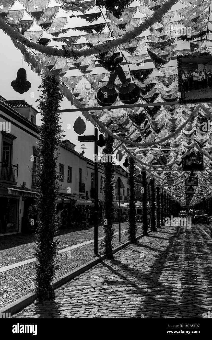 Algarve, Portugal – 2025.07.02: Eine kopfsteingepflasterte Straße mit leuchtenden Gelb-, Orange-, Schwarz- und Weißdekorationen für ein lokales Festival Stockfoto