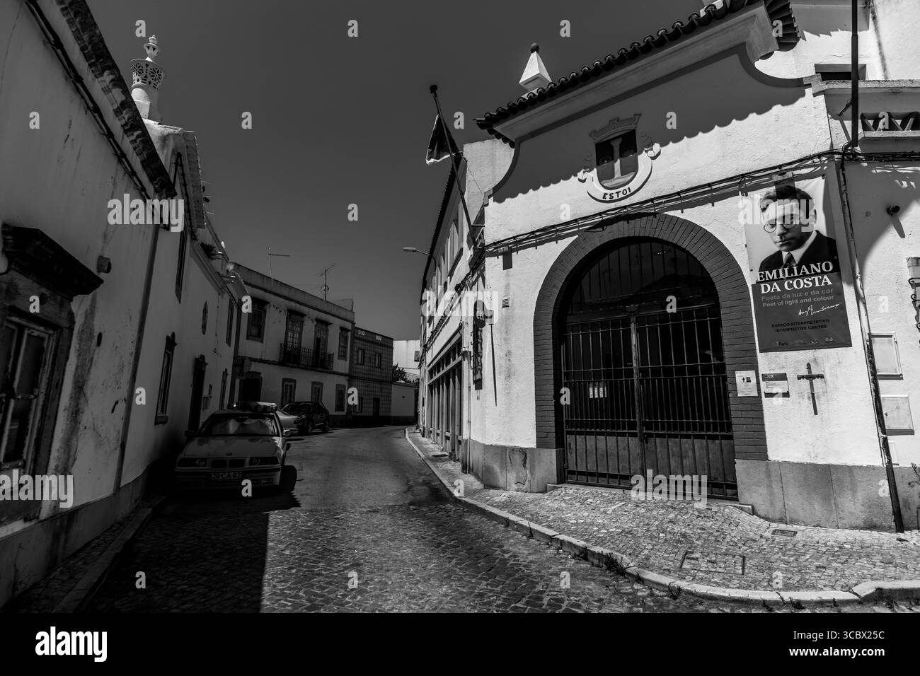Estoi, Algarve, Portugal – 2025.07.05: Ein historisches Marktgebäude mit einem Banner des Dichters Emiliano da Costa, Kopfsteinpflasterstraße und einem parkenden gelben Auto Stockfoto