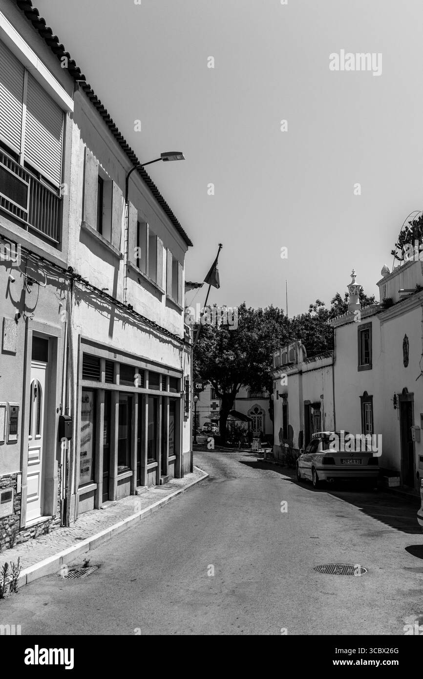 Estoi, Algarve, Portugal – 2025.07.05: Eine ruhige, enge Straße mit traditionellen Häusern, einem gelben BMW auf der Seite und dem portugiesischen fla Stockfoto