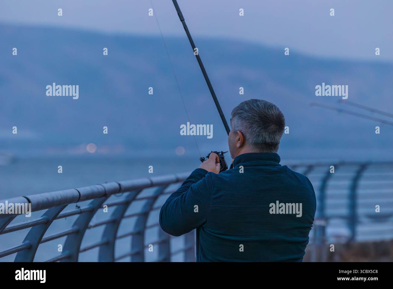 Rückansicht eines Menschen mit Angelrute am Pier am See von Galiläa in der Abenddämmerung, mit ruhigem Wasser und fernen Bergen Stockfoto