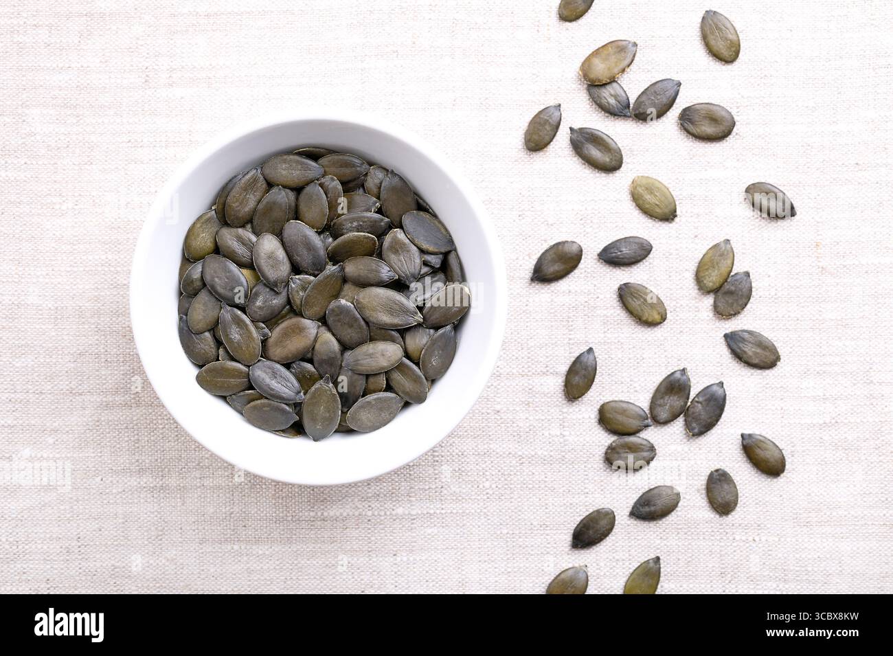 Steirische Ölkürbiskerne, in einer weißen Schüssel, auf Leinen. Getrocknete, dunkelgrüne und junge Früchte des Steirischen Kürbis, Cucurbita pepo var. Styriaca. Stockfoto