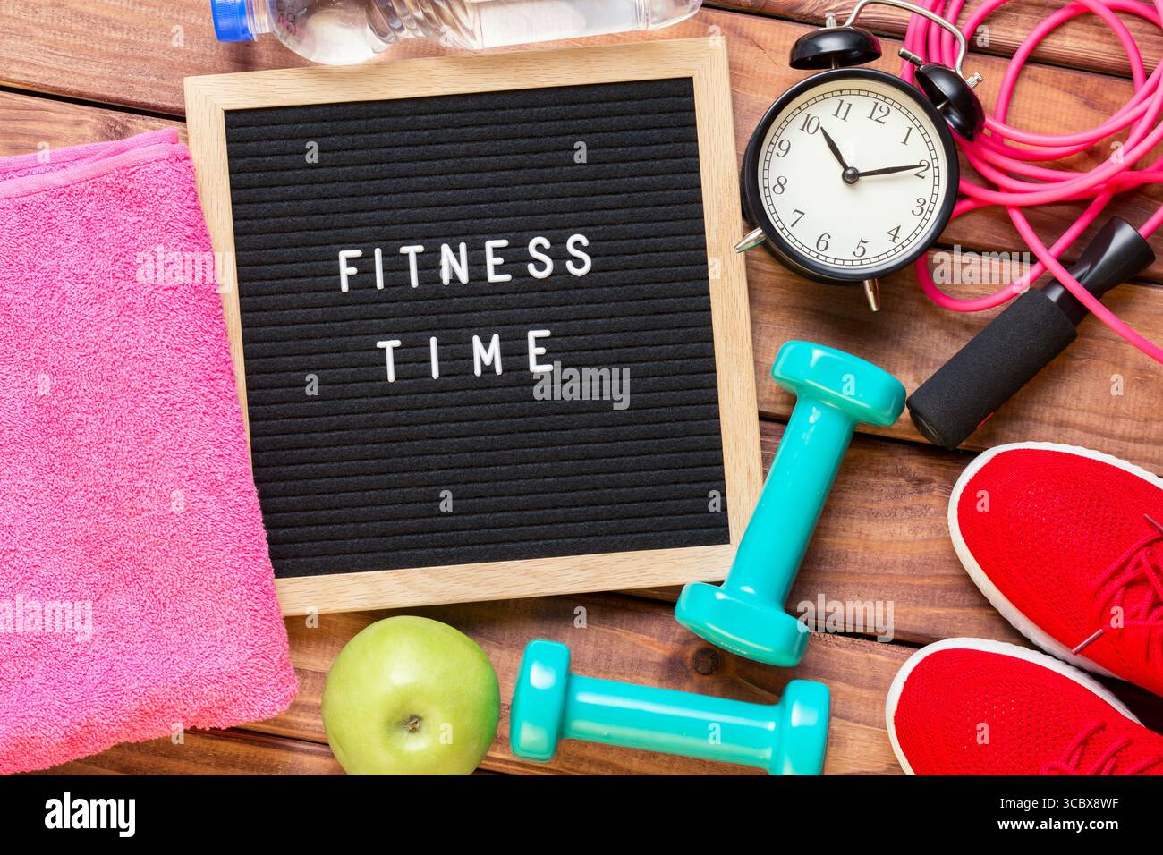 Fitnesszeit und Sportkonzept. Rote Turnschuhe, Apfel, Sprungseil, Kurzhanteln, rosa Handtuch, Flasche Wasser, Vintage-Wecker und schwarzes Schriftfeld mit Stockfoto