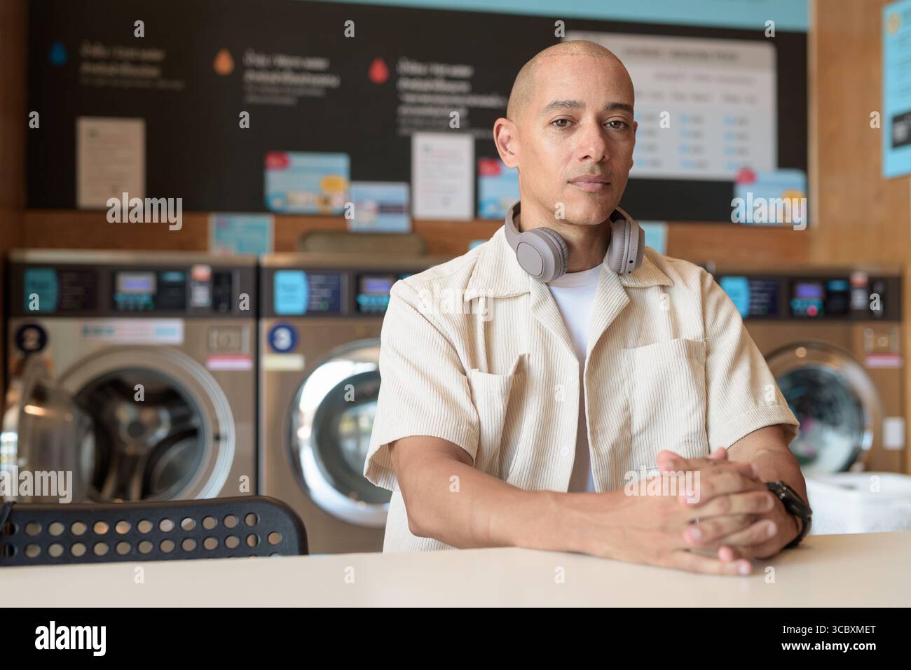 Kahler hispanischer Mann in lässigem Outfit, der in einem Selbstbedienungs-Wäschereiladen sitzt. Stadtporträt mit Waschmaschinen hinter ihm, zeigt alltägliche Stadtwelten Stockfoto