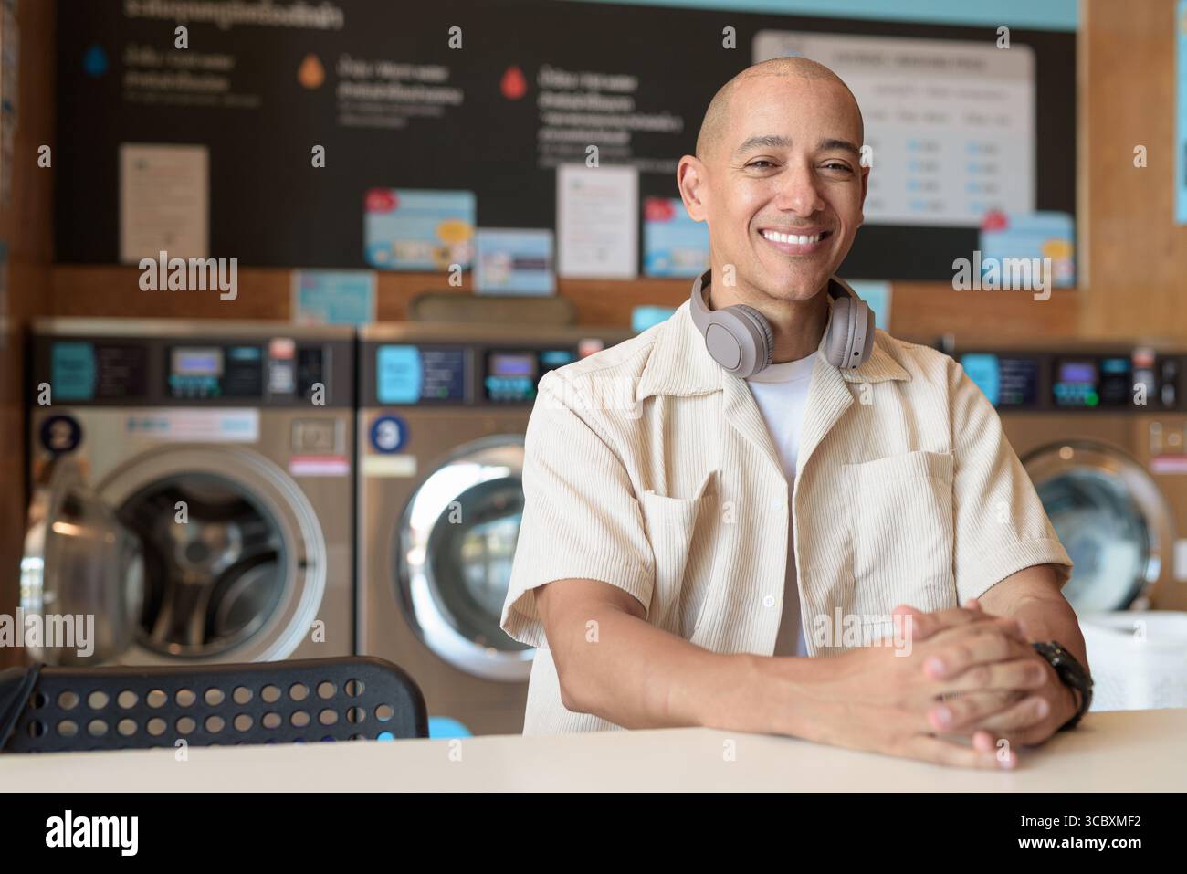 Kahler hispanischer Mann in lässigem Outfit, der in einem Selbstbedienungs-Wäschereiladen sitzt. Stadtporträt mit Waschmaschinen hinter ihm, zeigt alltägliche Stadtwelten Stockfoto