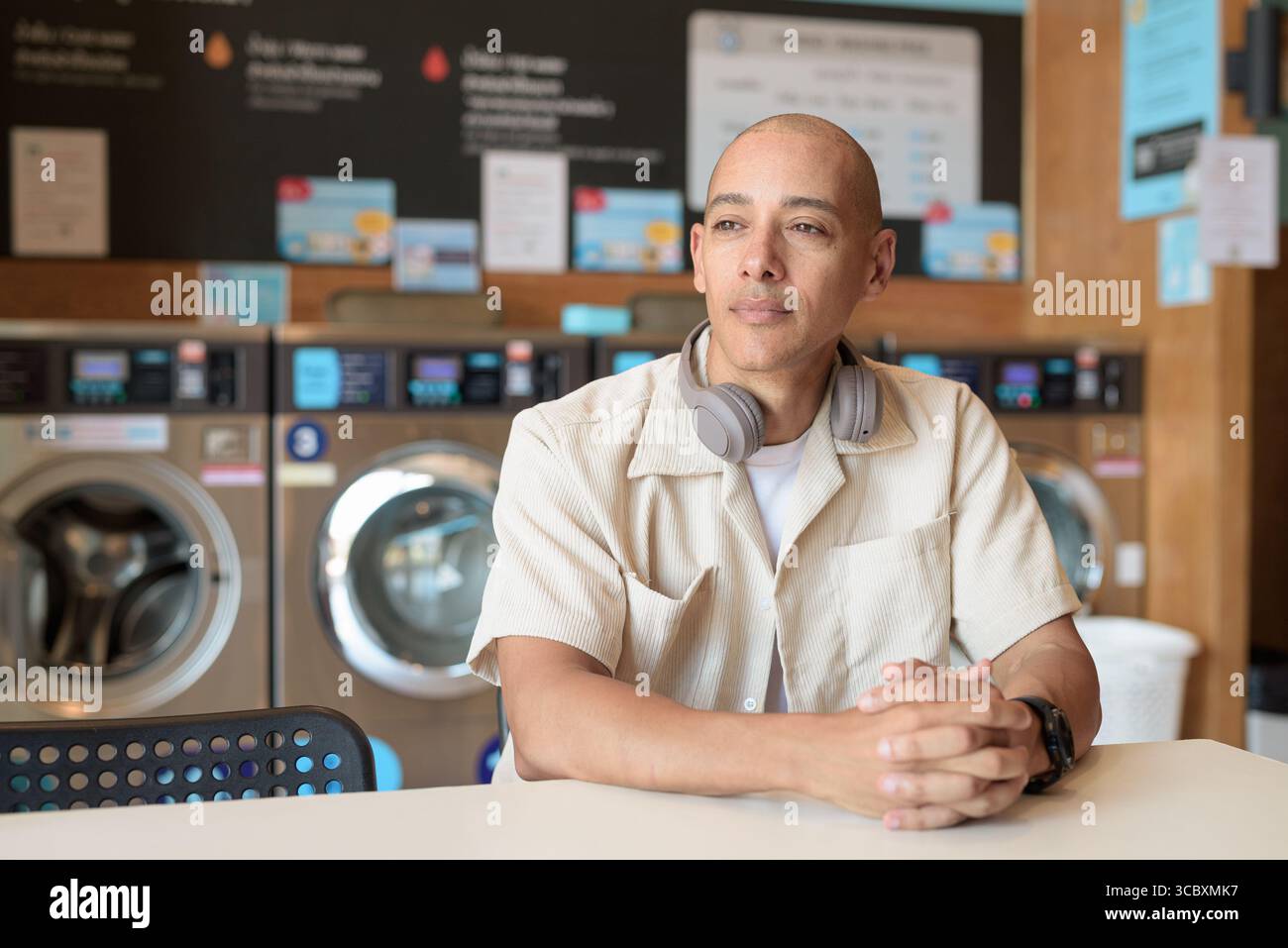 Kahler hispanischer Mann in lässigem Outfit, der in einem Selbstbedienungs-Wäschereiladen sitzt. Stadtporträt mit Waschmaschinen hinter ihm, zeigt alltägliche Stadtwelten Stockfoto