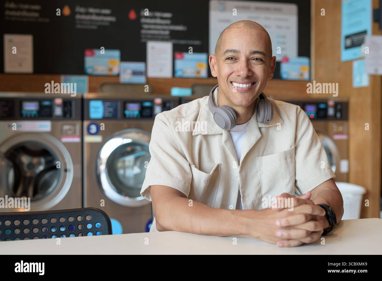 Kahler hispanischer Mann in lässigem Outfit, der in einem Selbstbedienungs-Wäschereiladen sitzt. Stadtporträt mit Waschmaschinen hinter ihm, zeigt alltägliche Stadtwelten Stockfoto