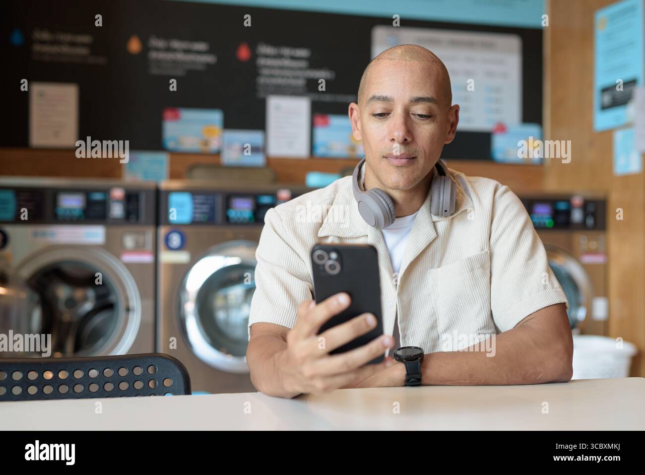 Kahler hispanischer Mann in legeren Kleidern, der in einer Selbstbedienungswäscherei mit Smartphone sitzt. Urbaner Alltag mit Waschmaschinen im Hintergrund Stockfoto