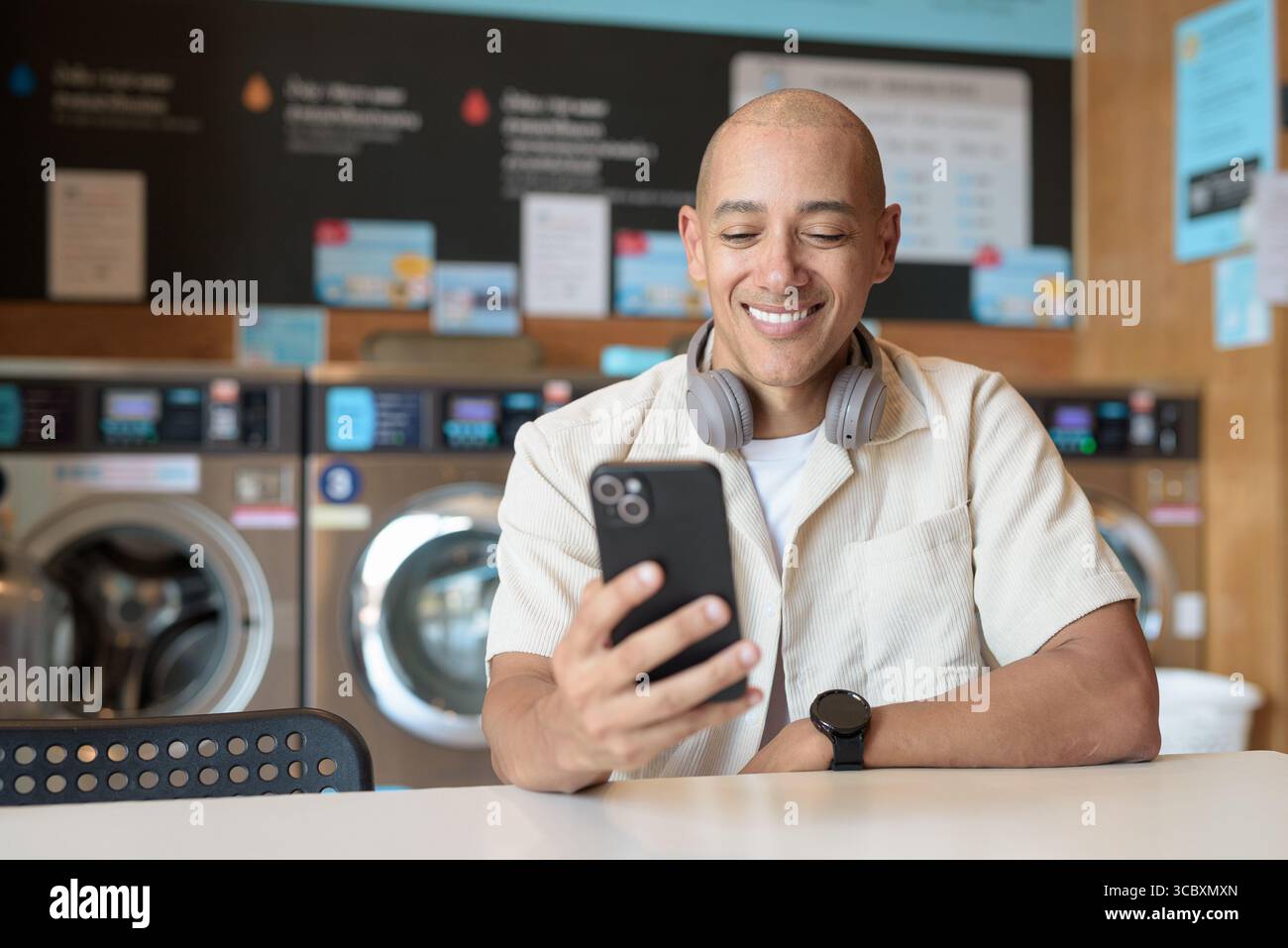 Kahler hispanischer Mann in legeren Kleidern, der in einer Selbstbedienungswäscherei mit Smartphone sitzt. Urbaner Alltag mit Waschmaschinen im Hintergrund Stockfoto