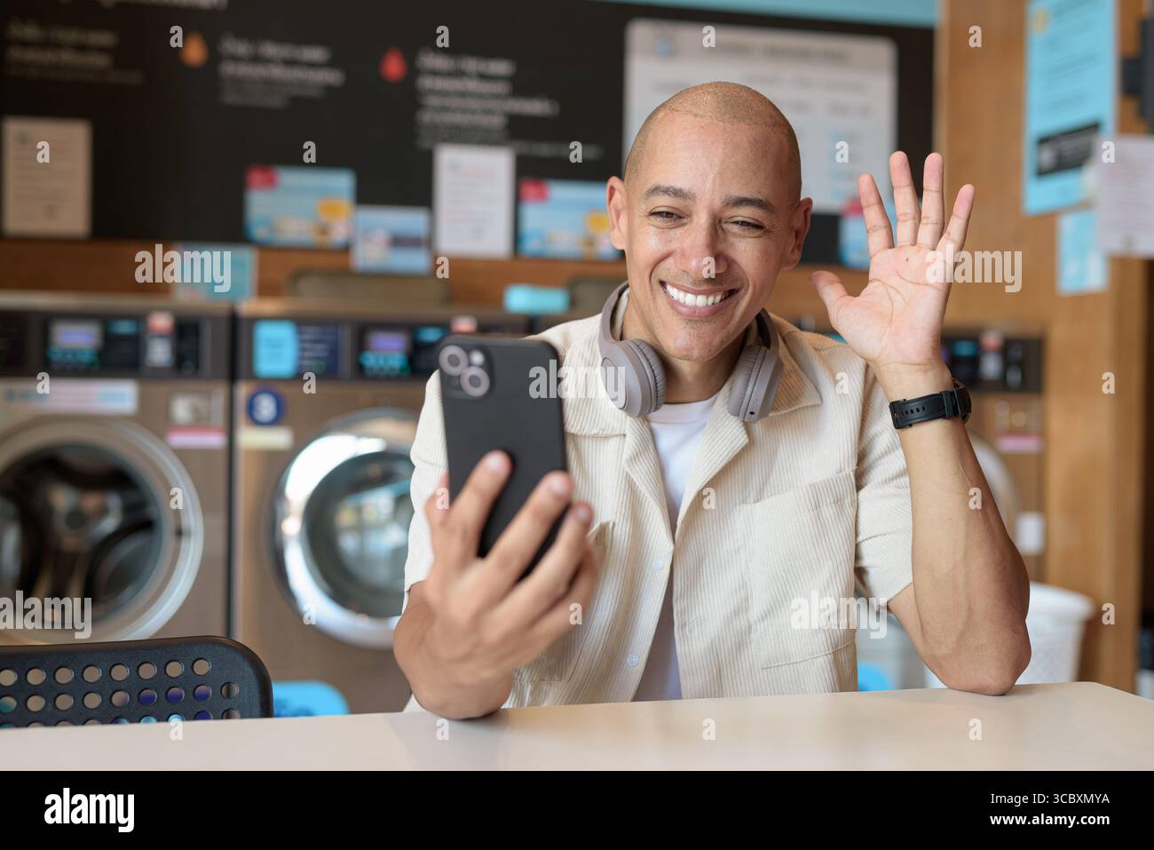 Kahler hispanischer Mann in legeren Kleidern, der in einer Selbstbedienungswäscherei mit Smartphone sitzt. Urbaner Alltag mit Waschmaschinen im Hintergrund Stockfoto