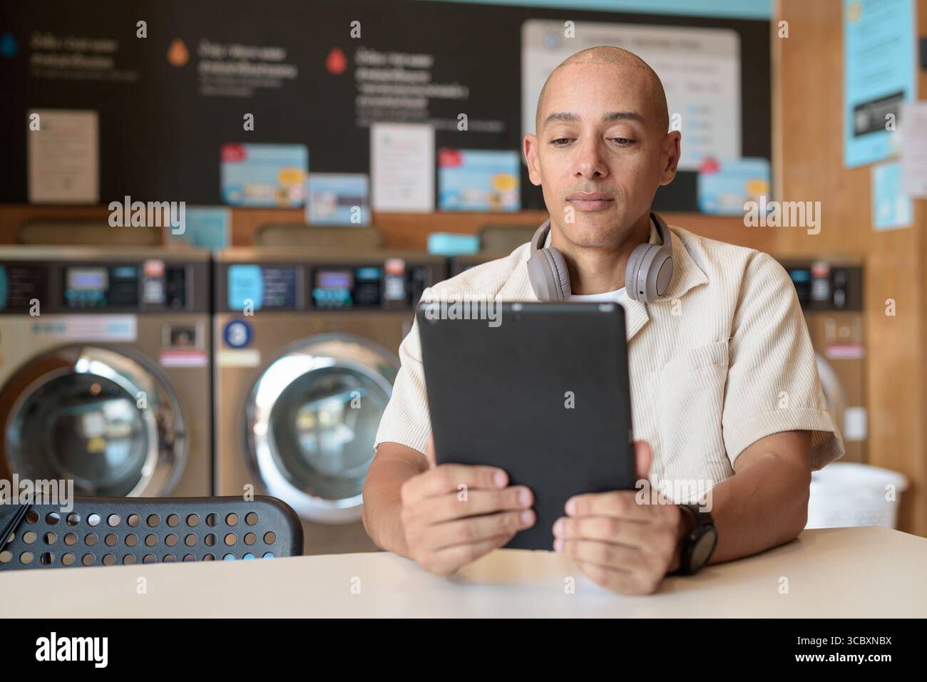 Kahler hispanischer Mann in lässiger Kleidung, der in einer Wäscherei sitzt und ein digitales Tablet benutzt. Urbanes Alltagskonzept mit Waschmaschinen im Hintergrund Stockfoto