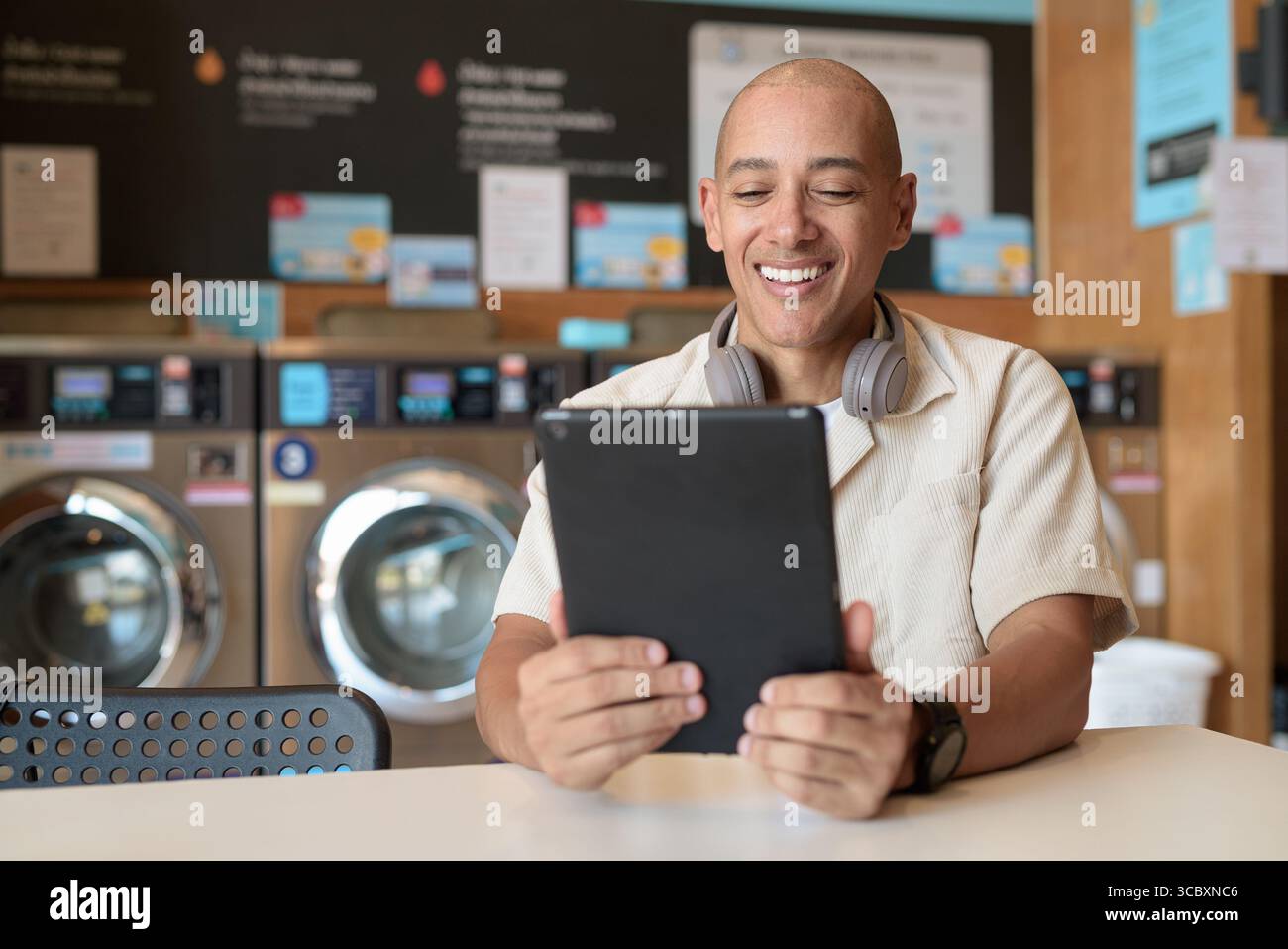 Kahler hispanischer Mann in lässiger Kleidung, der in einer Wäscherei sitzt und ein digitales Tablet benutzt. Urbanes Alltagskonzept mit Waschmaschinen im Hintergrund Stockfoto