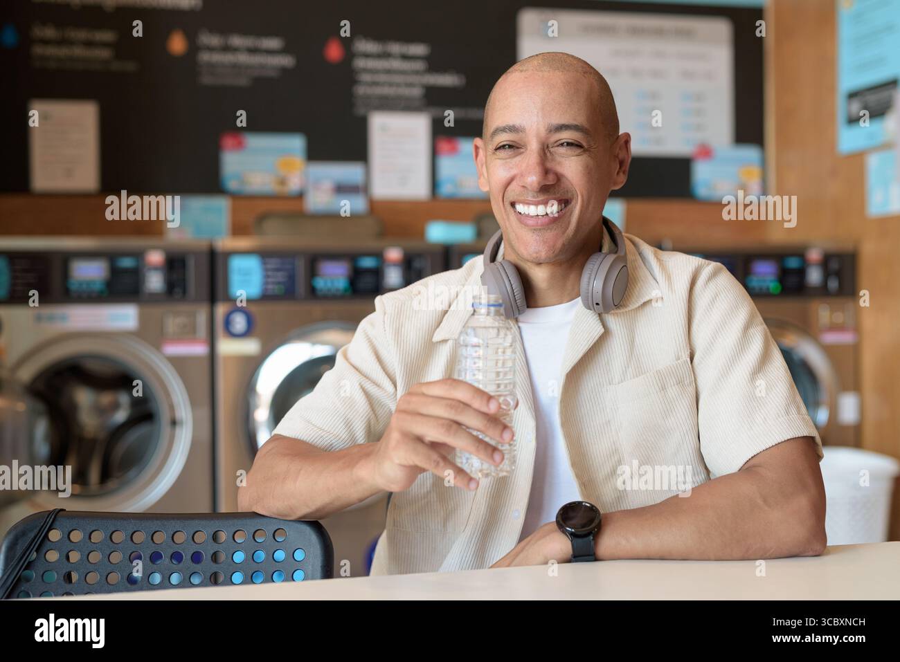 Kahler hispanischer Mann in lässigem Outfit, der in einem Wäschereiladen sitzt und aus einer Wasserflasche trinkt. Urbaner Alltag mit Waschmaschinen im Hintergrund. Stockfoto