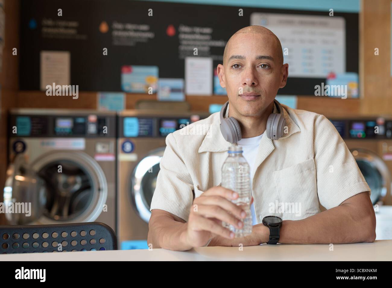 Kahler hispanischer Mann in lässigem Outfit, der in einem Wäschereiladen sitzt und aus einer Wasserflasche trinkt. Urbaner Alltag mit Waschmaschinen im Hintergrund. Stockfoto
