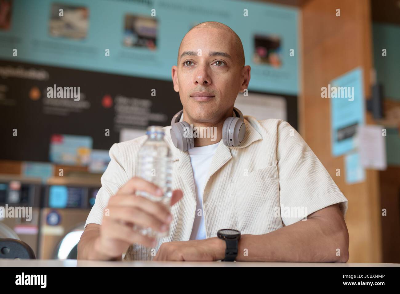 Kahler hispanischer Mann in lässigem Outfit, der in einem Wäschereiladen sitzt und aus einer Wasserflasche trinkt. Urbaner Alltag mit Waschmaschinen im Hintergrund. Stockfoto