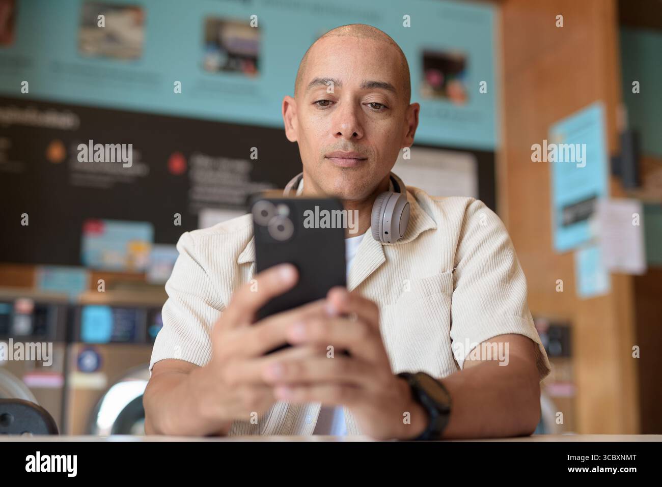 Kahler hispanischer Mann in legeren Kleidern, der in einer Selbstbedienungswäscherei mit Smartphone sitzt. Urbaner Alltag mit Waschmaschinen im Hintergrund Stockfoto