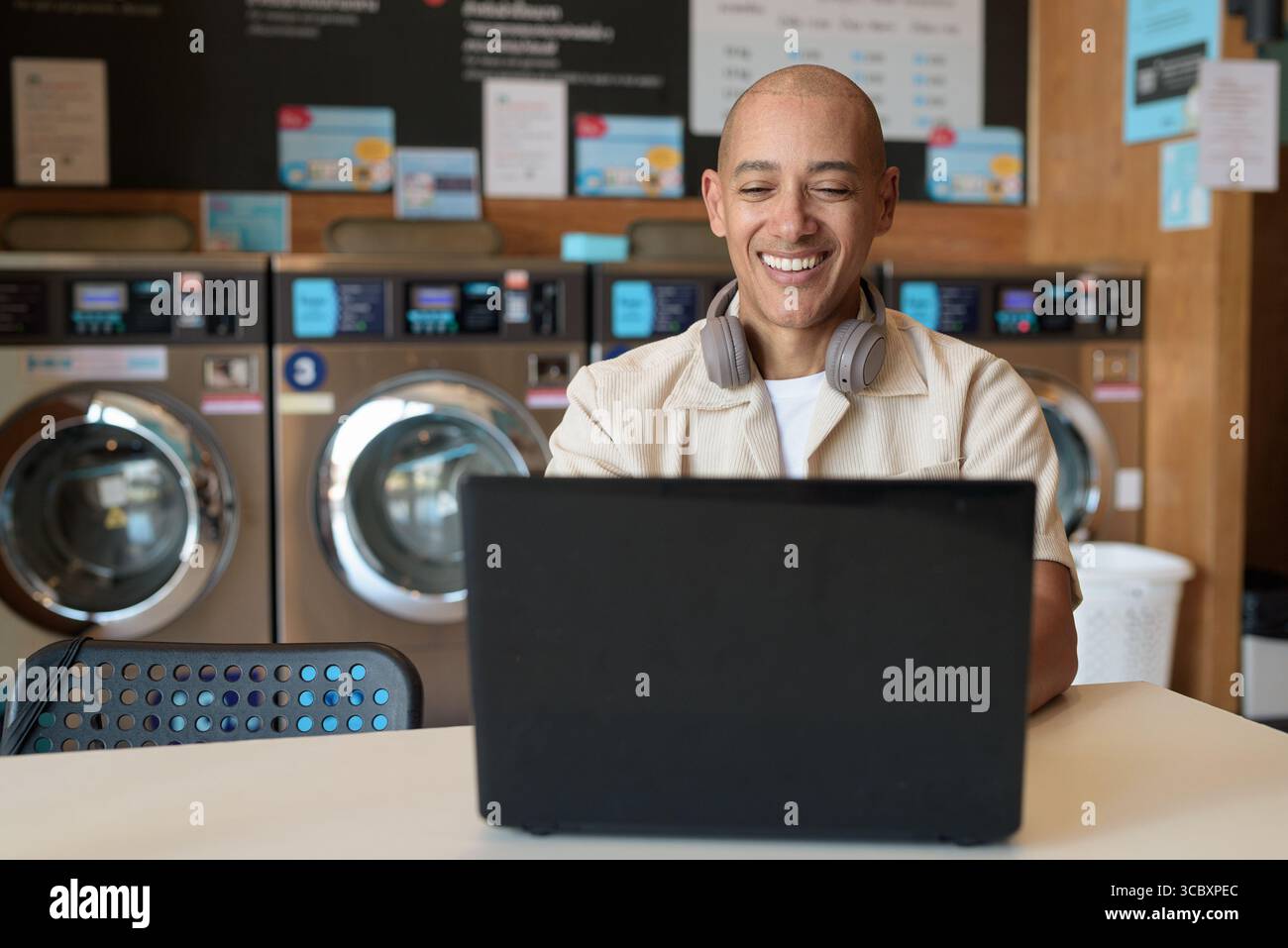 Kahler hispanischer Mann in legeren Kleidern, der in einem Wäscheshop sitzt und einen Laptop benutzt. Urbanes Alltagskonzept mit Waschmaschinen im Hintergrund Stockfoto