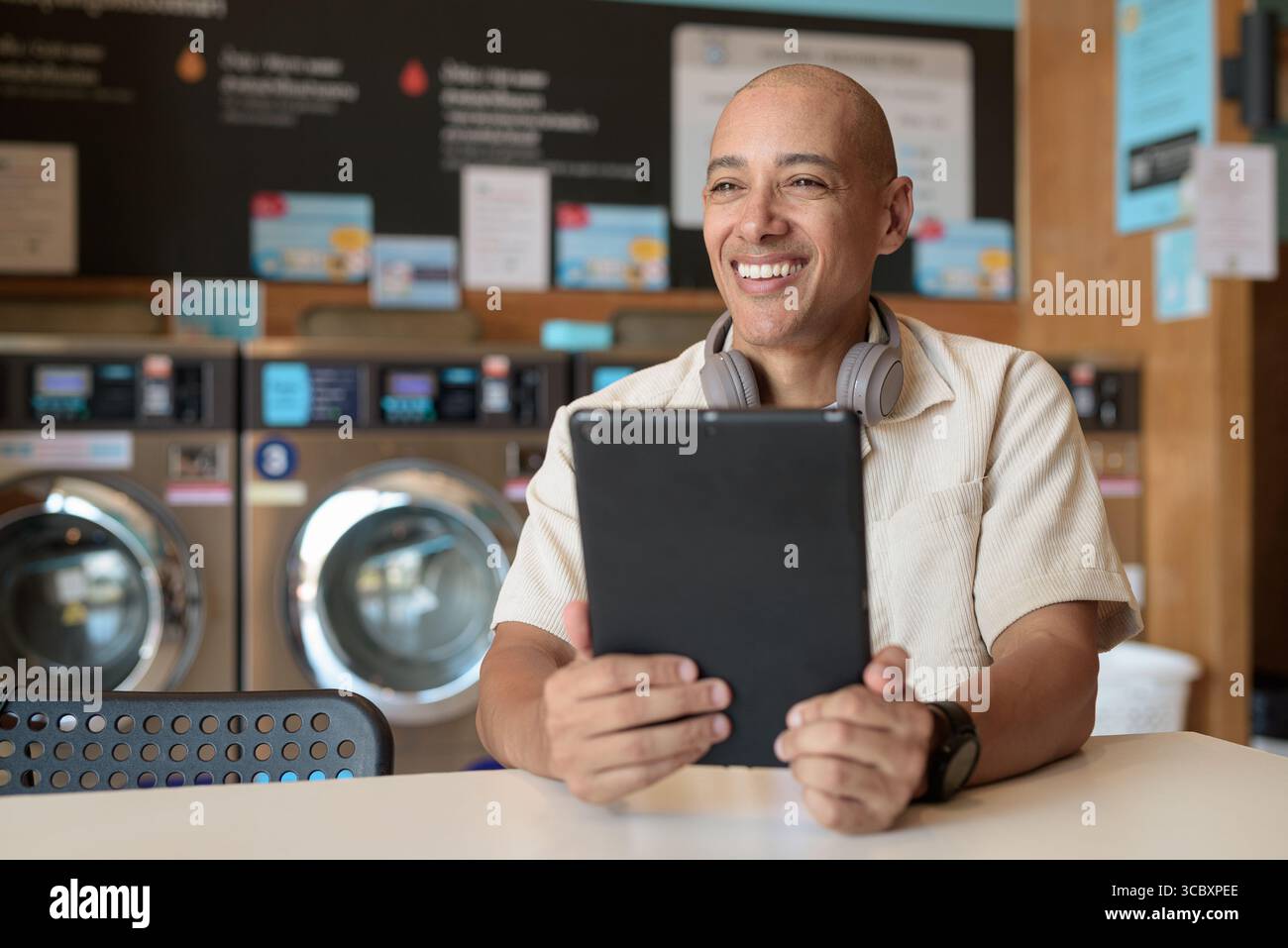 Kahler hispanischer Mann in lässiger Kleidung, der in einer Wäscherei sitzt und ein digitales Tablet benutzt. Urbanes Alltagskonzept mit Waschmaschinen im Hintergrund Stockfoto